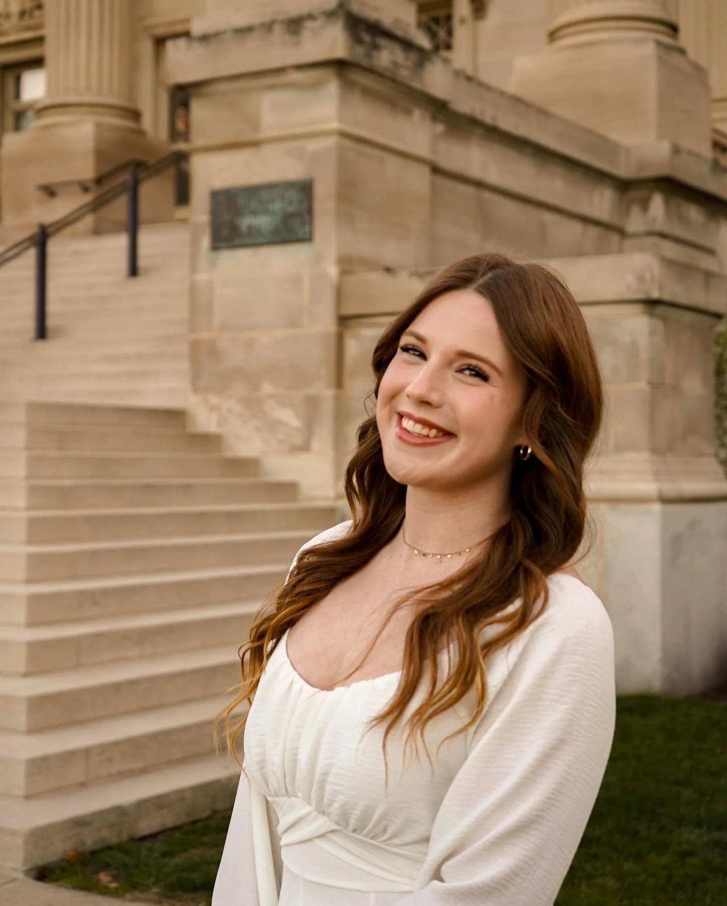 A young woman with long, wavy brown hair and light skin, smiling in front of a stone building with steps.