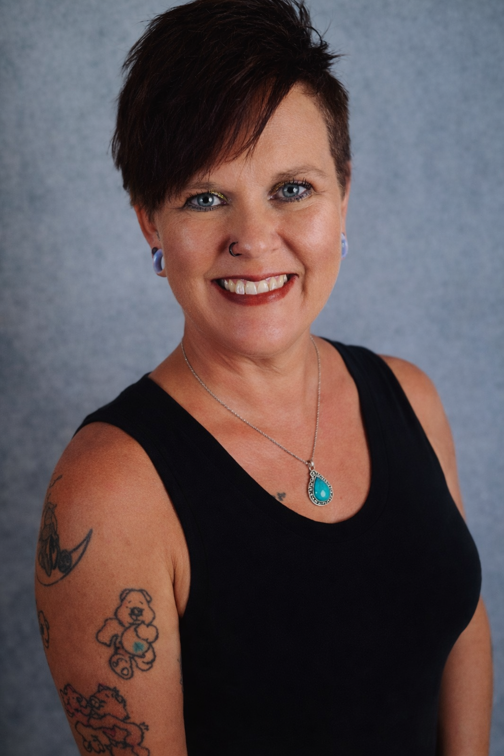 A woman with short, edgy dark hair with red highlights, smiling, wearing a sleeveless olive green top, with tattoos on her left arm, and a nose ring, standing against a gray background.