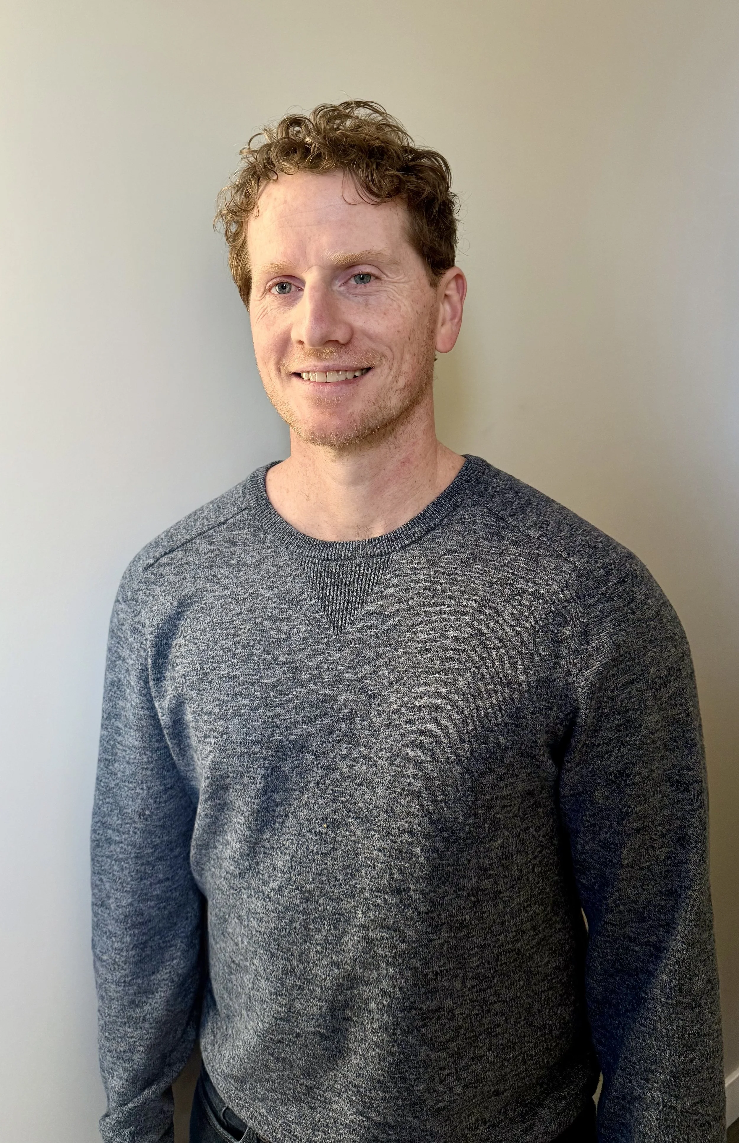 A man with curly hair and light skin, smiling, wearing a gray sweater, standing against a plain light-colored wall.