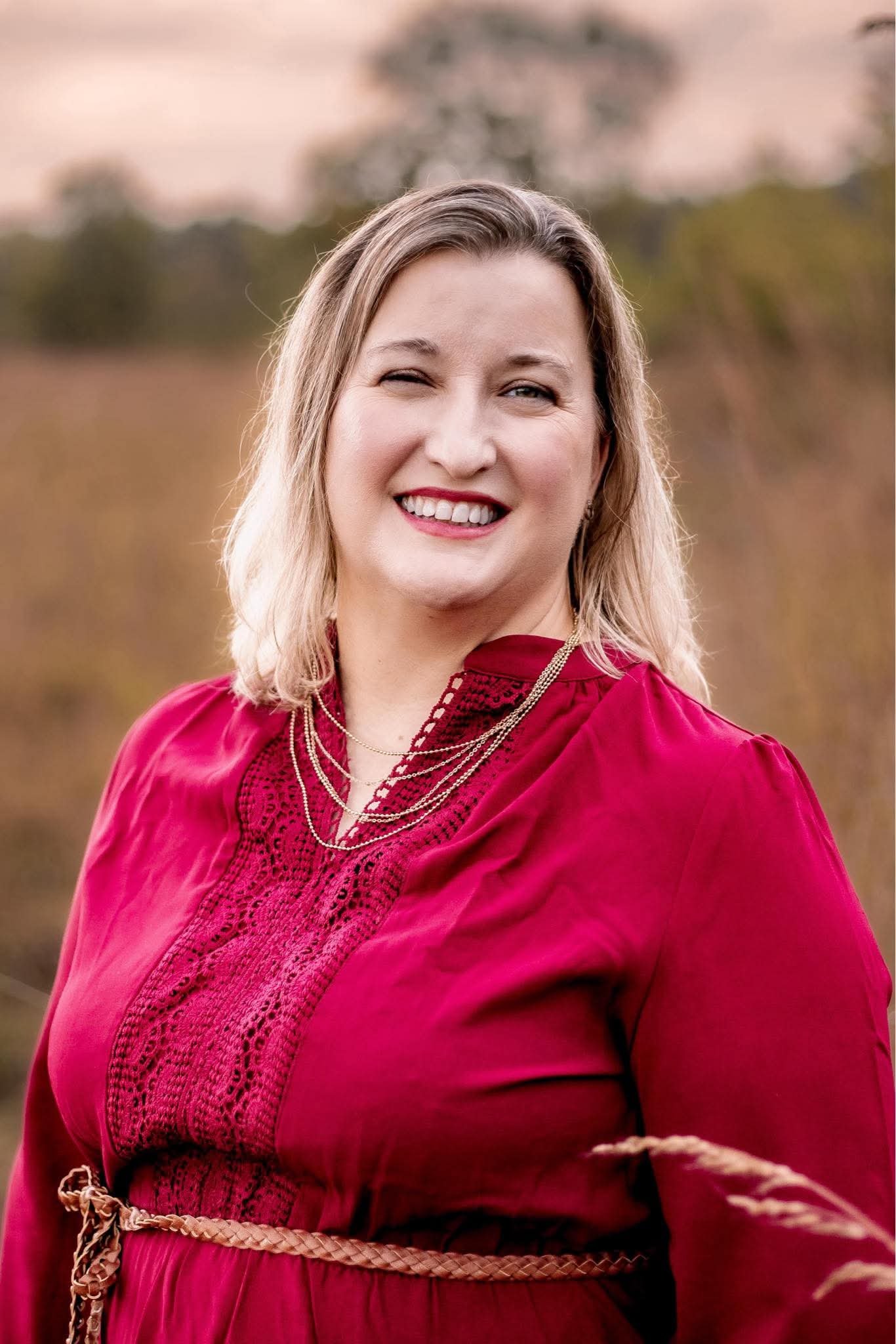 A smiling woman with blonde hair in a red dress outdoors during sunset or golden hour.