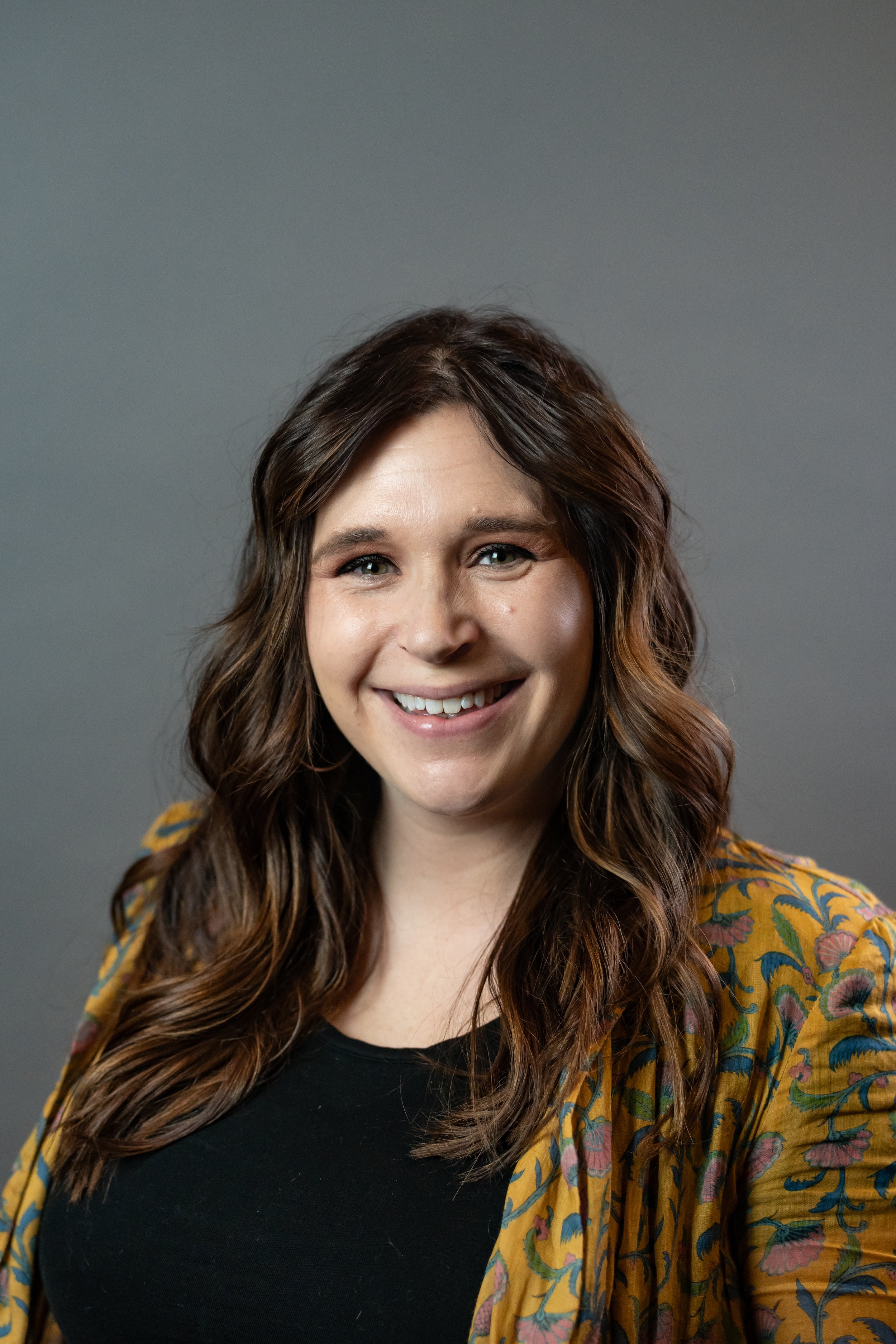 Headshot of a woman with long, wavy brown hair, smiling, wearing a black top and a yellow floral blazer against a gray background.
