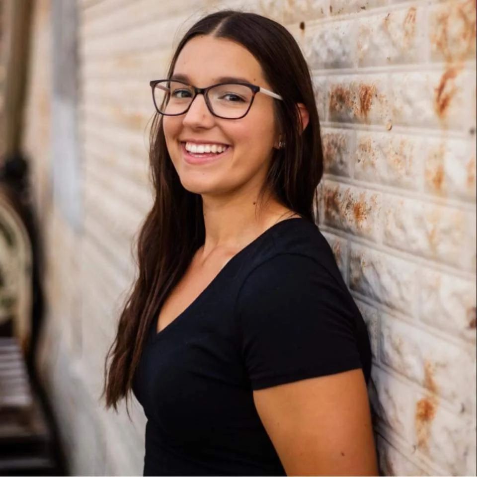 Young woman with long brown hair, wearing glasses and a black t-shirt, smiling and leaning against a textured brick wall.