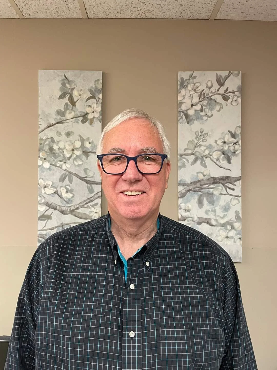 A smiling man with white hair and glasses wearing a black checkered shirt standing in front of two paintings of blossoming branches on a beige wall.