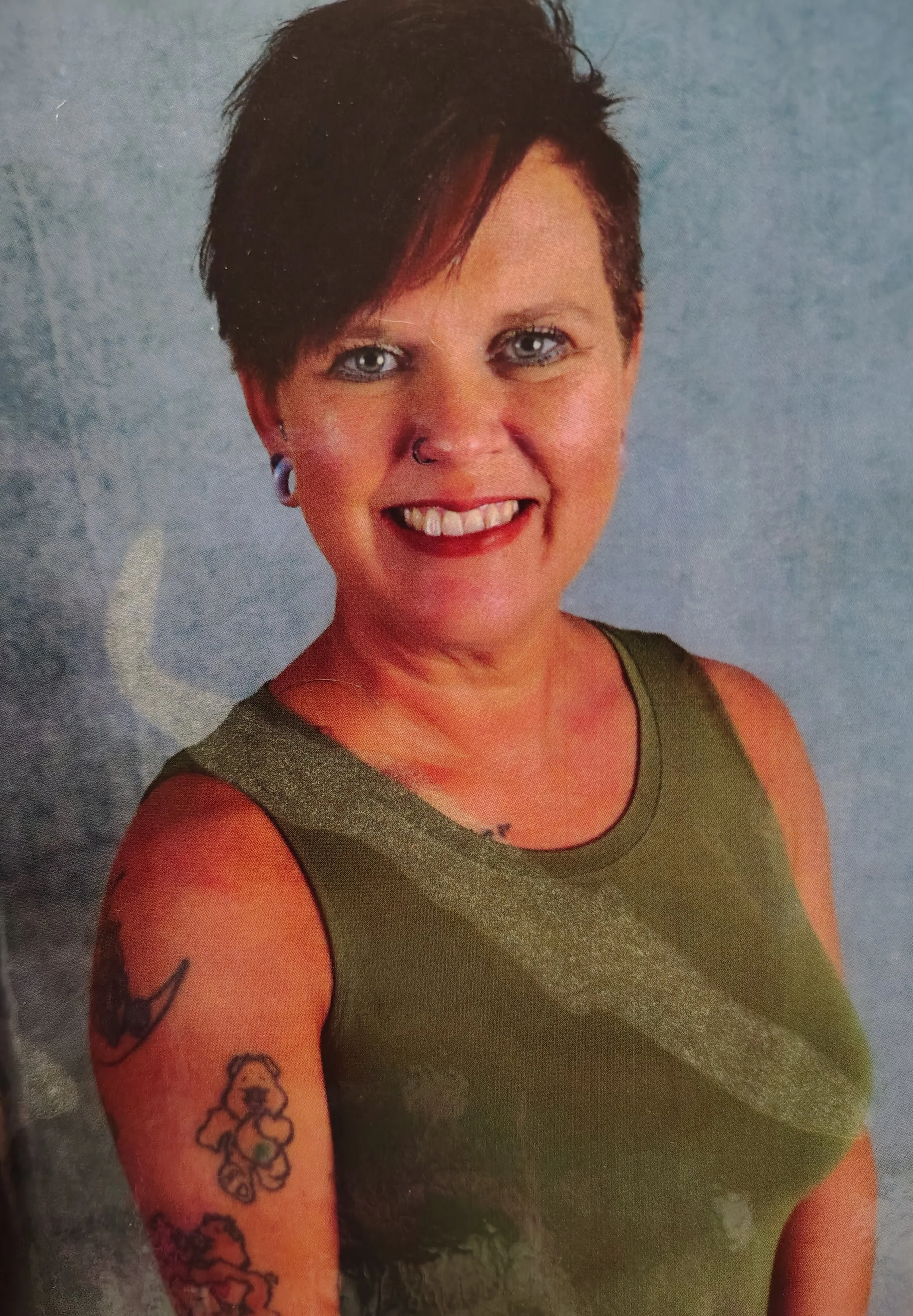 A woman with short, edgy dark hair with red highlights, smiling, wearing a sleeveless olive green top, with tattoos on her left arm, and a nose ring, standing against a gray background.
