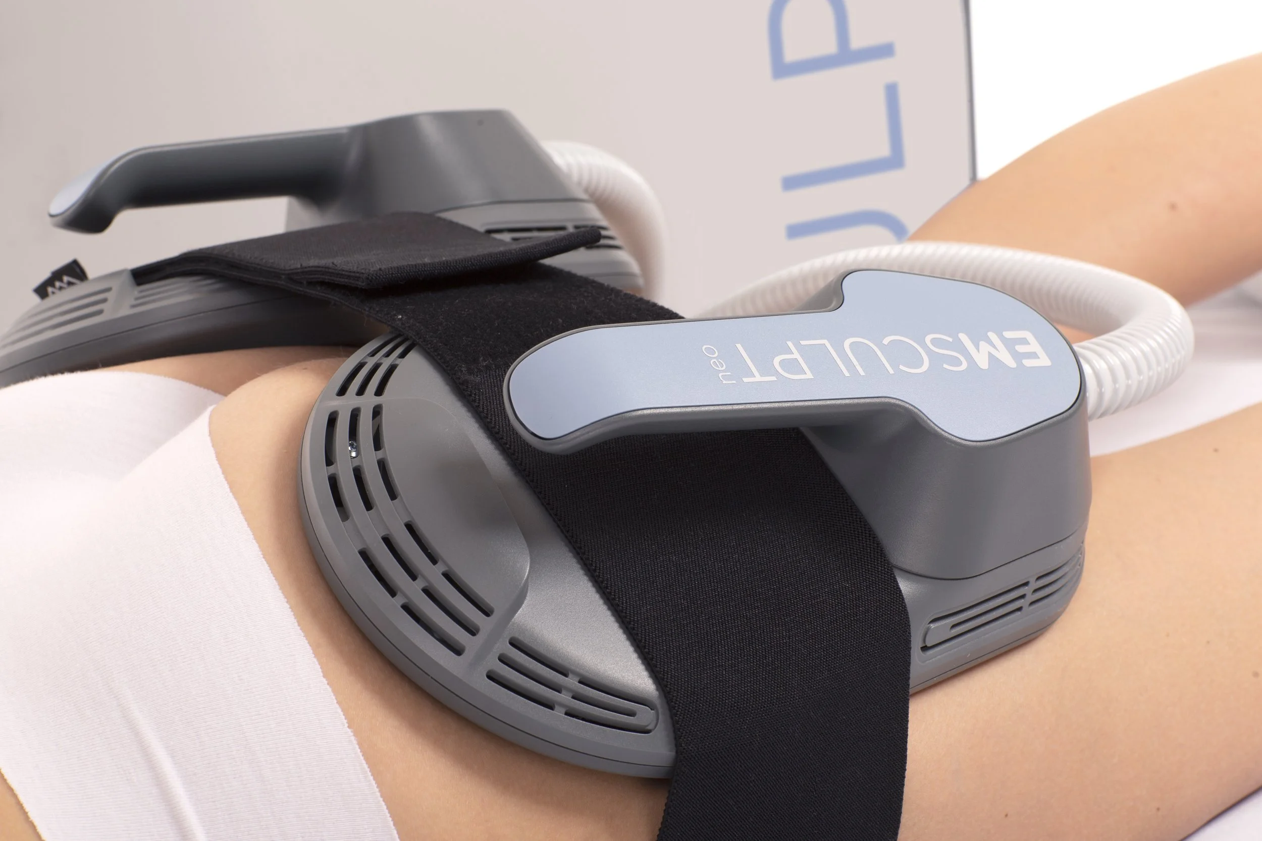 Medical device, EmSculpt Neo, attached to a person's abdomen with black straps, used for body contouring or muscle building.
