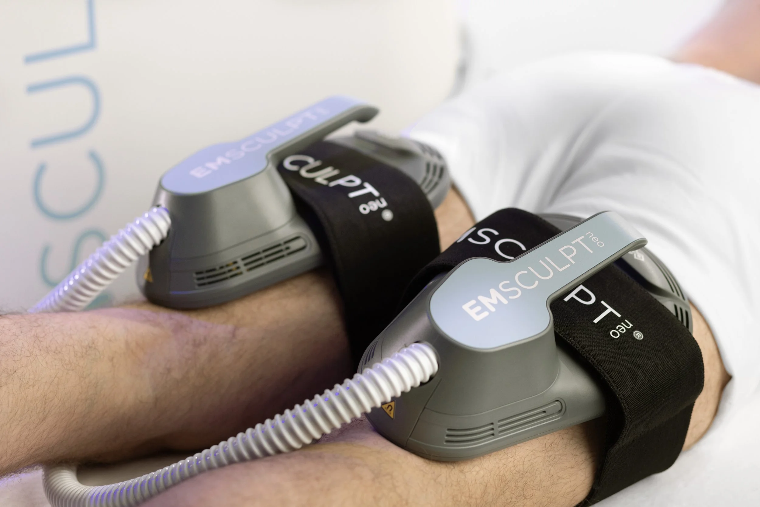 Close-up of a person's arm with two EM Sculpt Neo devices strapped with black bands, connected by white cords, likely used for body contouring or muscle toning.