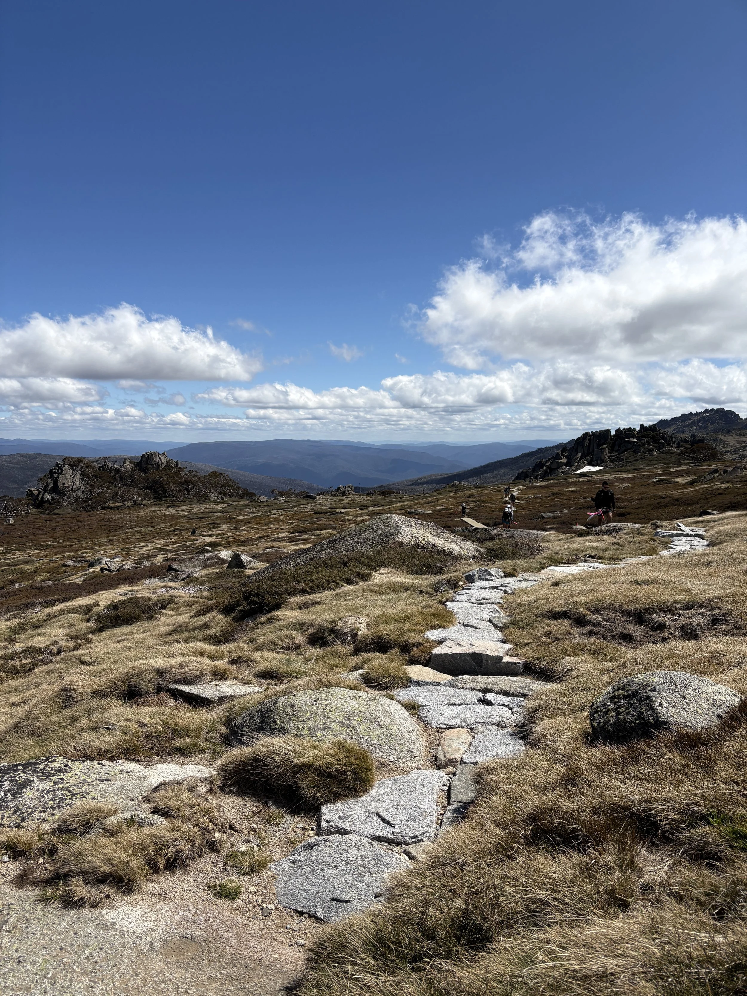 A mountain landscape featuring a rocky trail, dry grass, and a few hikers walking along the path under a partly cloudy sky with rolling mountains in the distance.