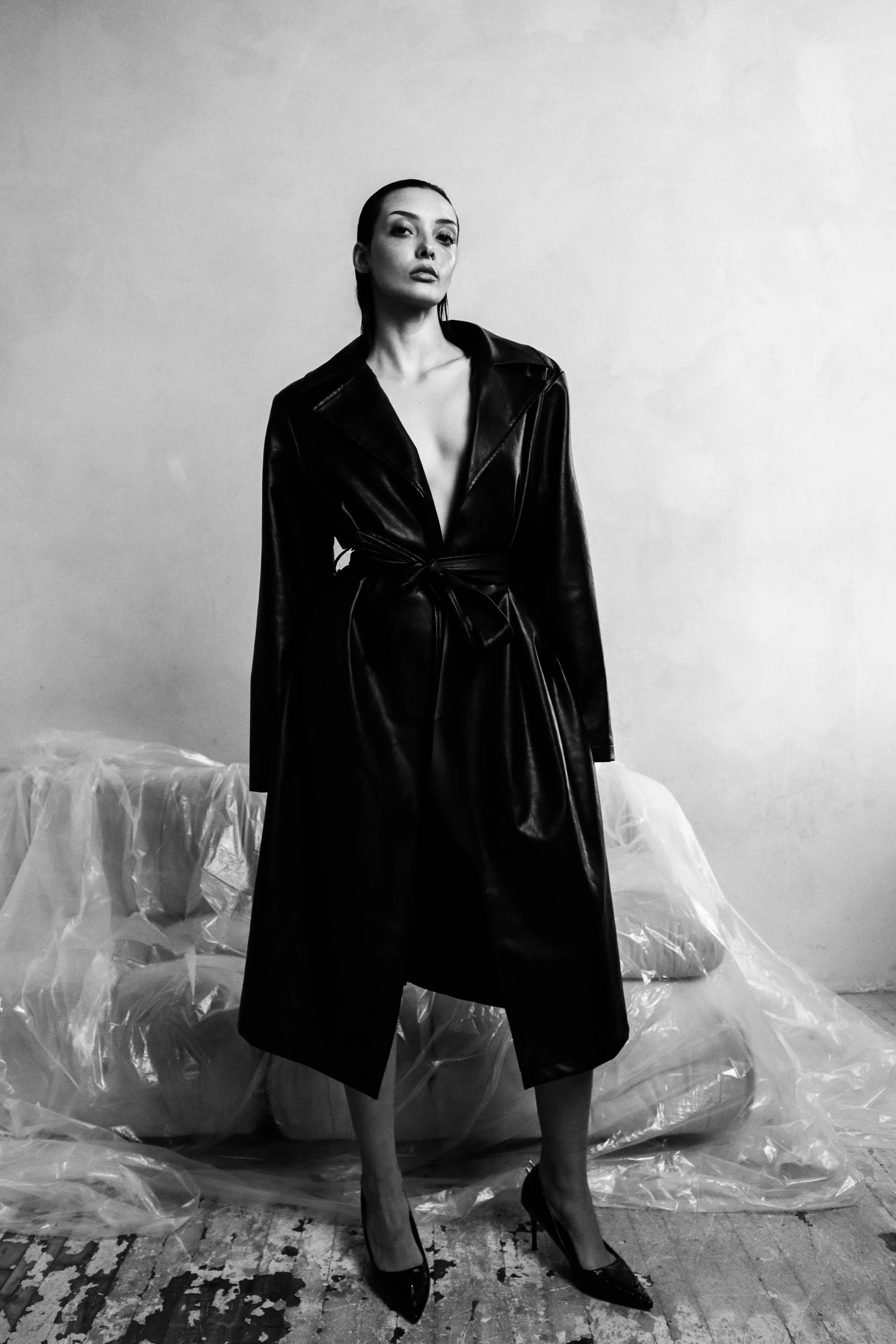A woman wearing a long, shiny black leather trench coat and high heels standing in front of a plain wall with a plastic-covered object behind her in a black and white photo.
