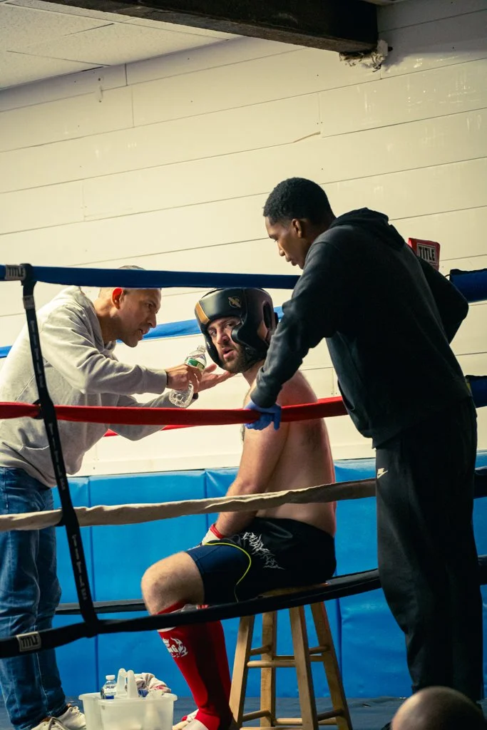 A boxer sitting on a stool in a boxing ring, receiving medical attention from two men, one holding a water bottle and the other adjusting his headgear.