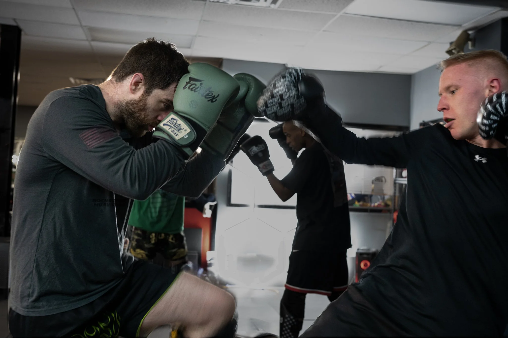Two men in boxing gloves training in a gym. One man is crouched with his hands up, preparing to block, while the other is throwing a punch.