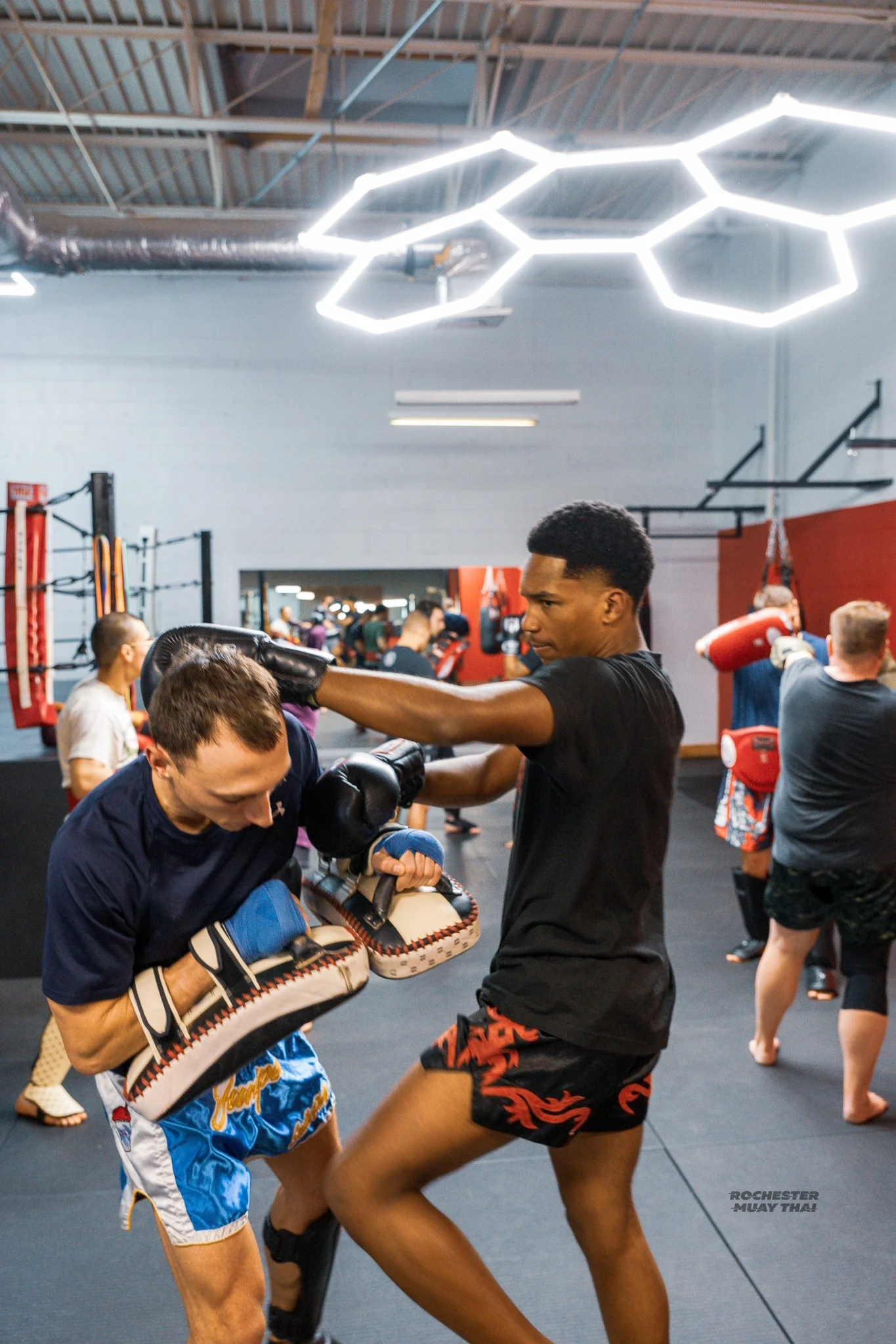 Two men practicing Muay Thai in a gym. The man on the left is throwing a punch, while the man on the right blocks with his arm. Several people are training in the background.