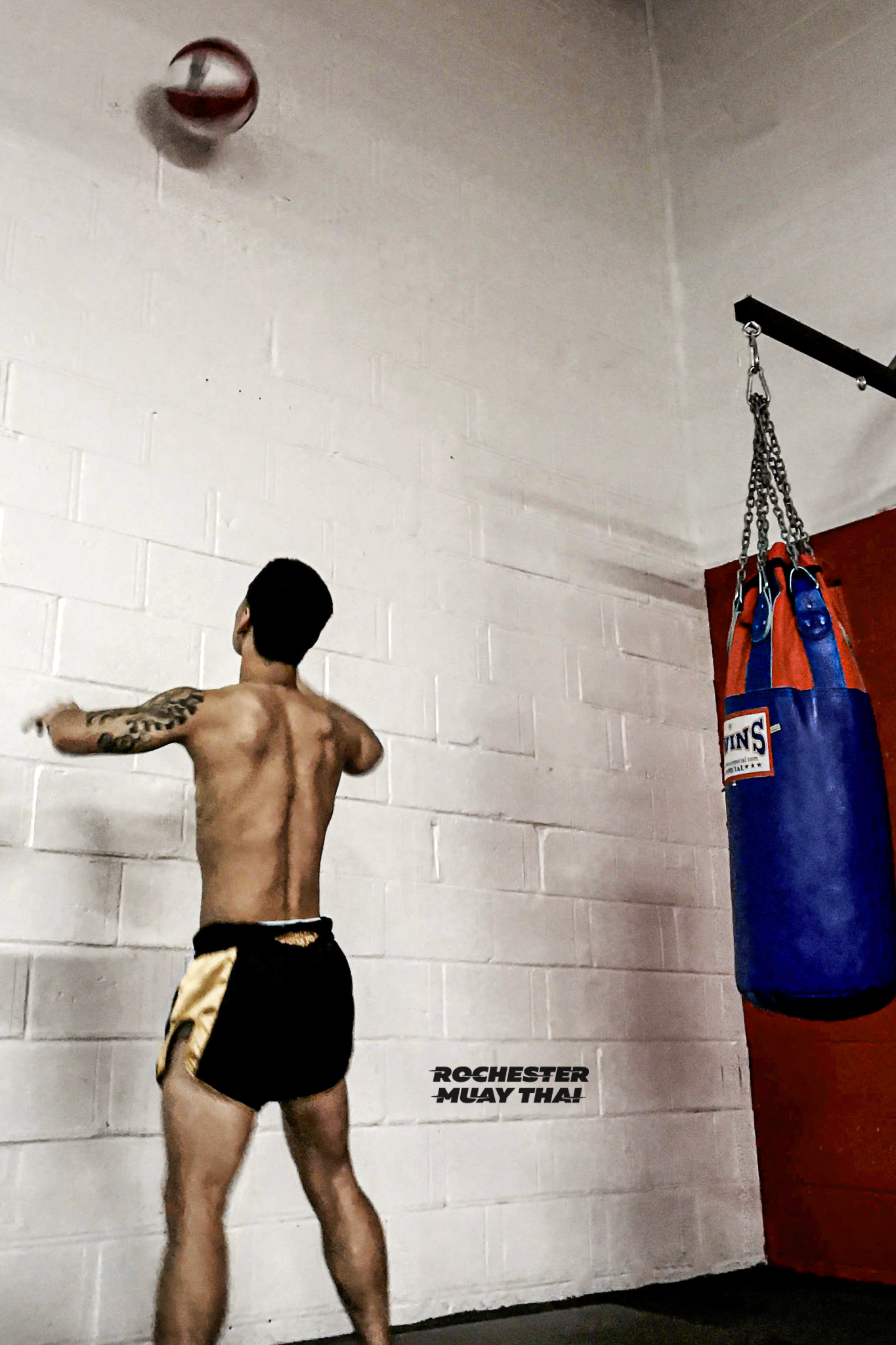 A shirtless male Muay Thai fighter in black and gold shorts practicing punches in a gym with a punching bag and the text 'ROCHESTER MUAY THAI' on the wall.