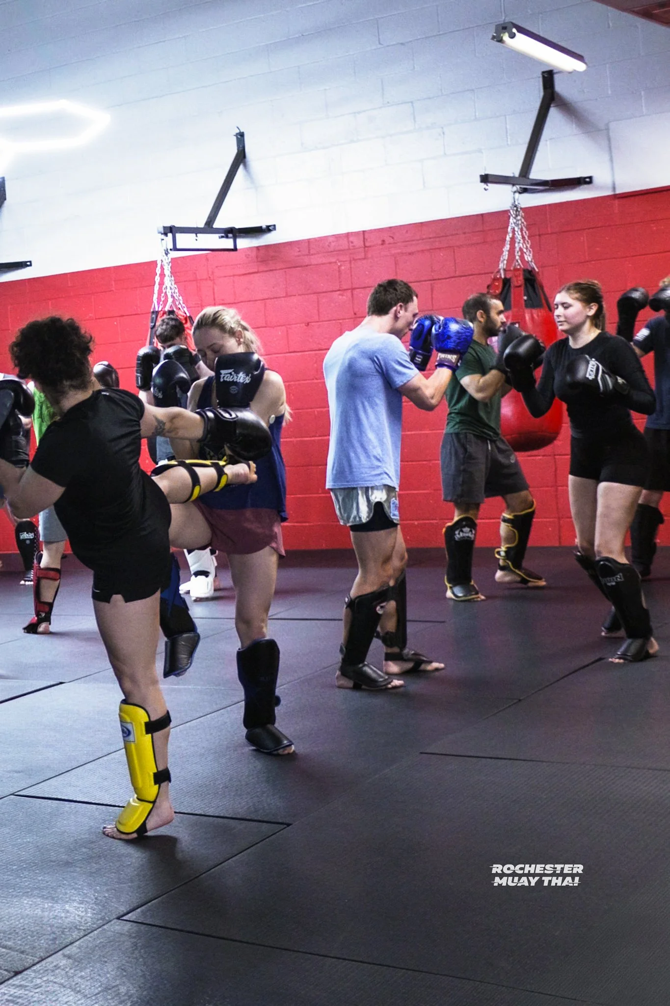 People practicing Muay Thai in a gym, wearing gloves and protective gear, with punching bags in the background.
