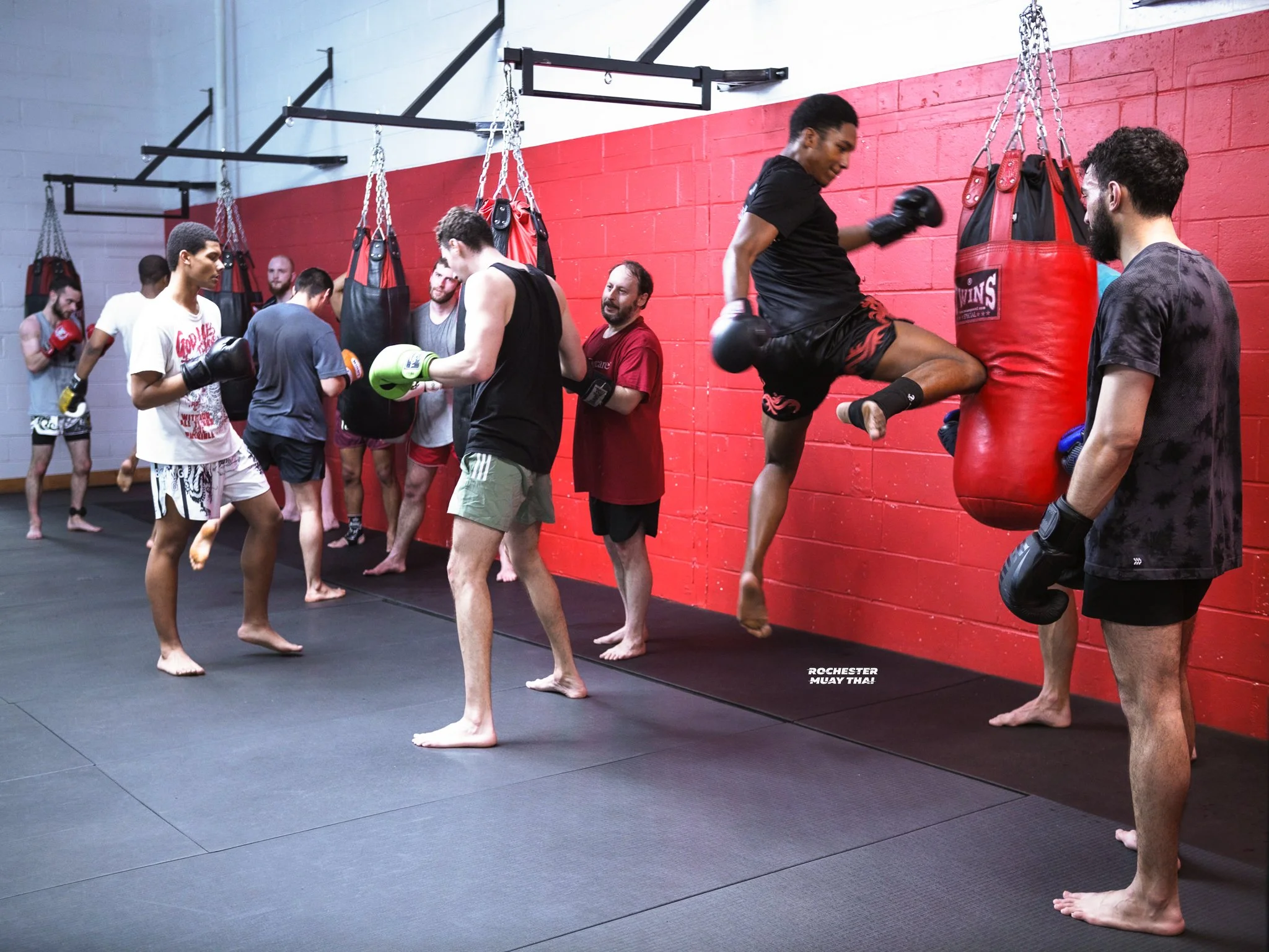 Group of people practicing martial arts in a gym, some using boxing gloves, others training kickboxing with punching bags, on a black padded floor with a red and gray wall in the background.