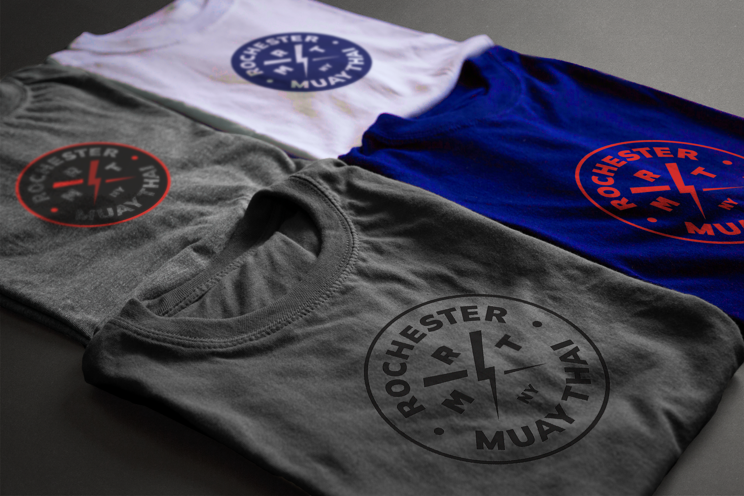 Three t-shirts in white, gray, and blue, each with a round logo for Rochester Muay Thai, featuring a lightning bolt and text.