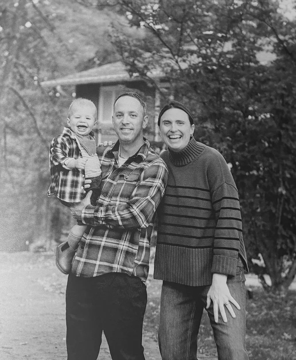 A black-and-white photo of a family of three outside in a wooded area. The man is holding a child, and they are all smiling at the camera. The woman standing next to them is also smiling.