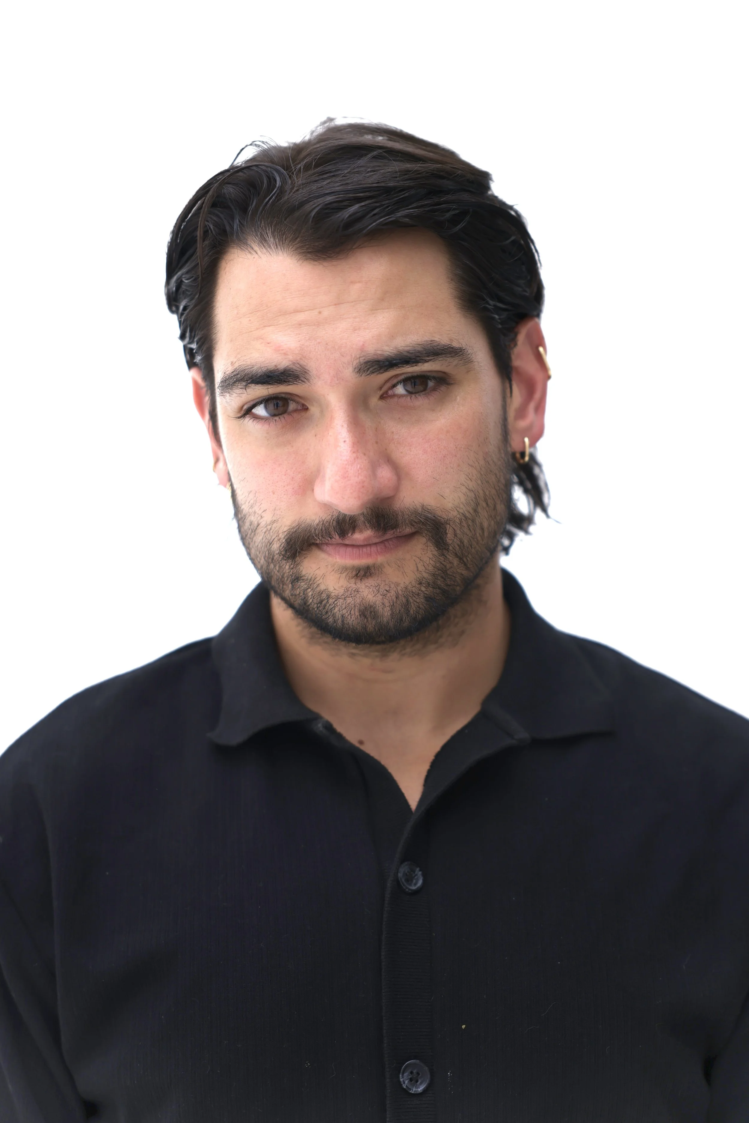Close-up of a young man with dark hair, beard, and earrings, wearing a black button-up shirt against a white background.