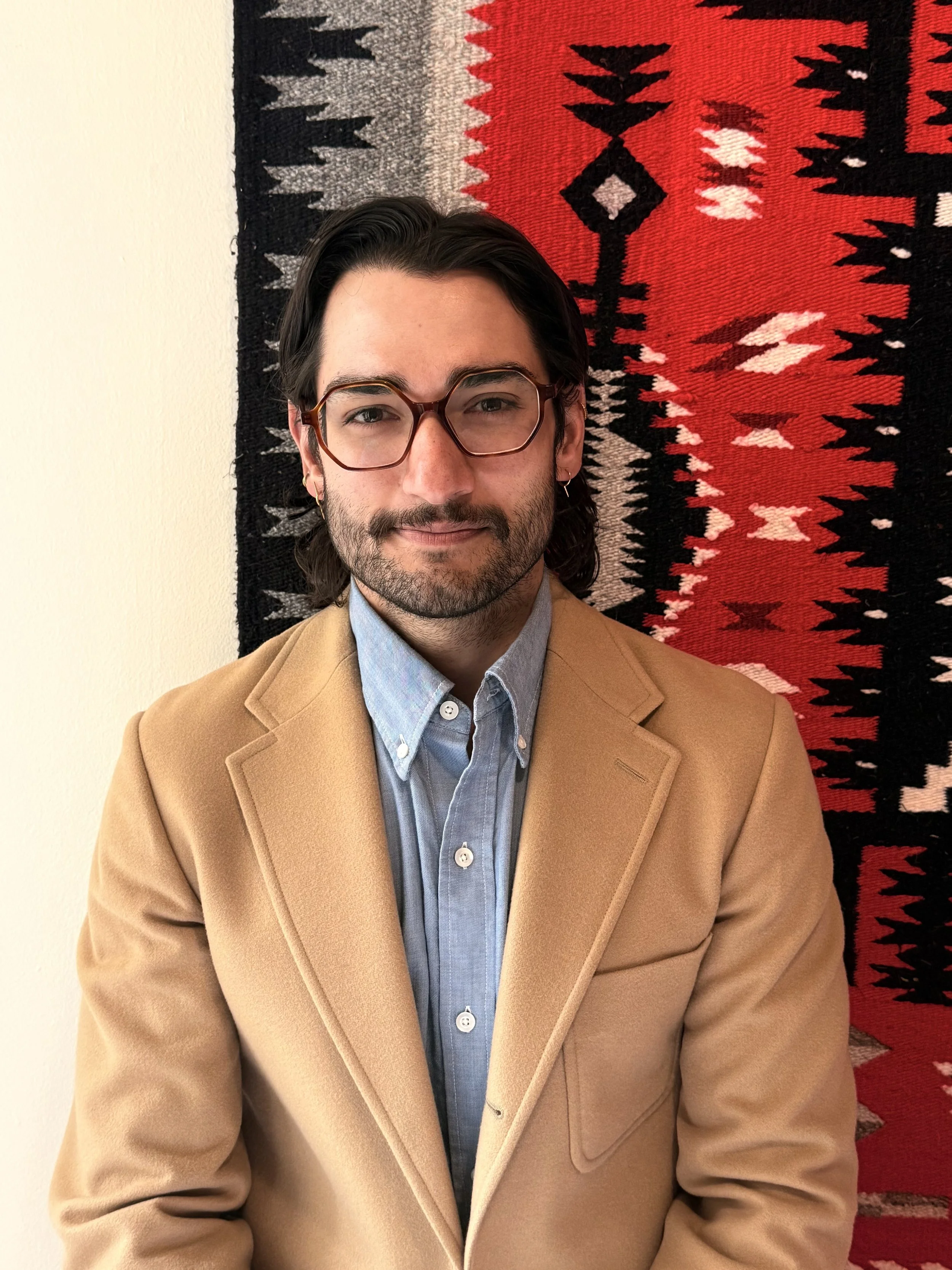 Close-up of a young man with dark hair, glasses, and a beard, wearing a blue shirt and a tan blazer, standing in front of a red, black, gray, and white woven textile wall hanging.