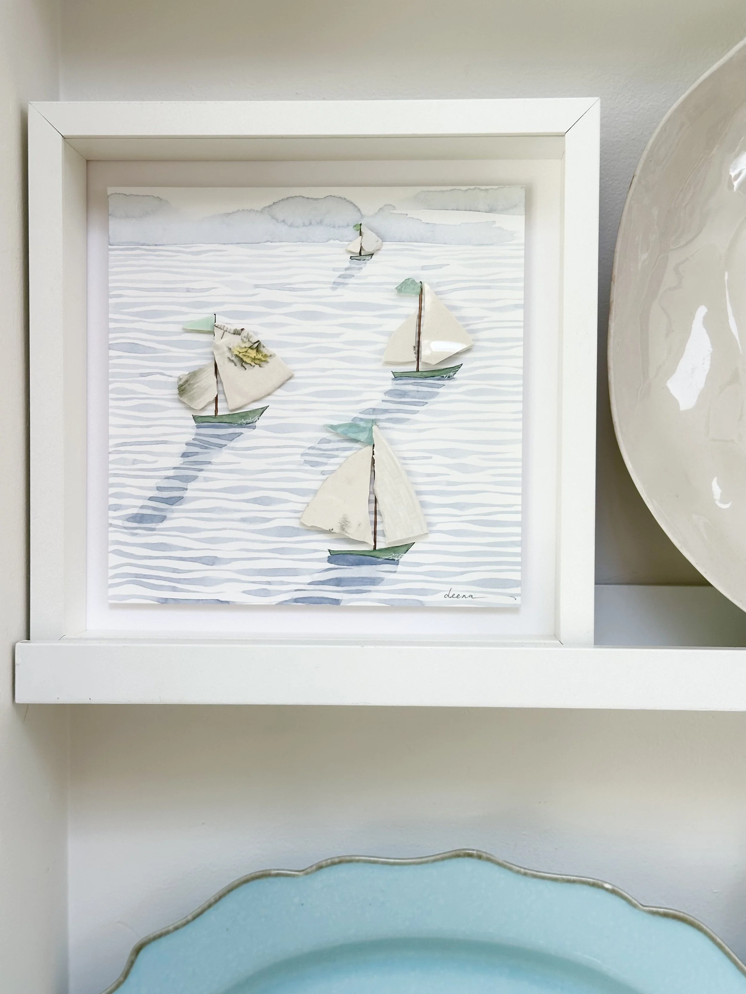 Mixed media coastal artwork with watercolour waves and sailboats made from reclaimed china shards in a white frame.