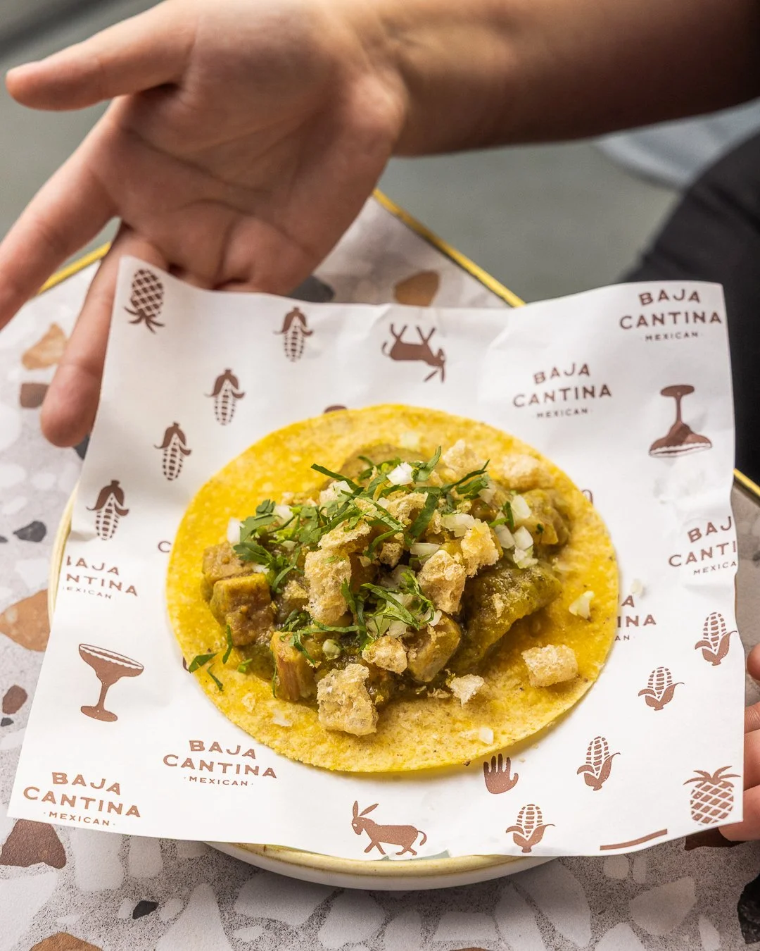 A taco with a yellow corn shell filled with meat, topped with chopped onions and cilantro, on paper printed with Mexican motifs and the text 'Baja Cantina' and 'Mexican'.