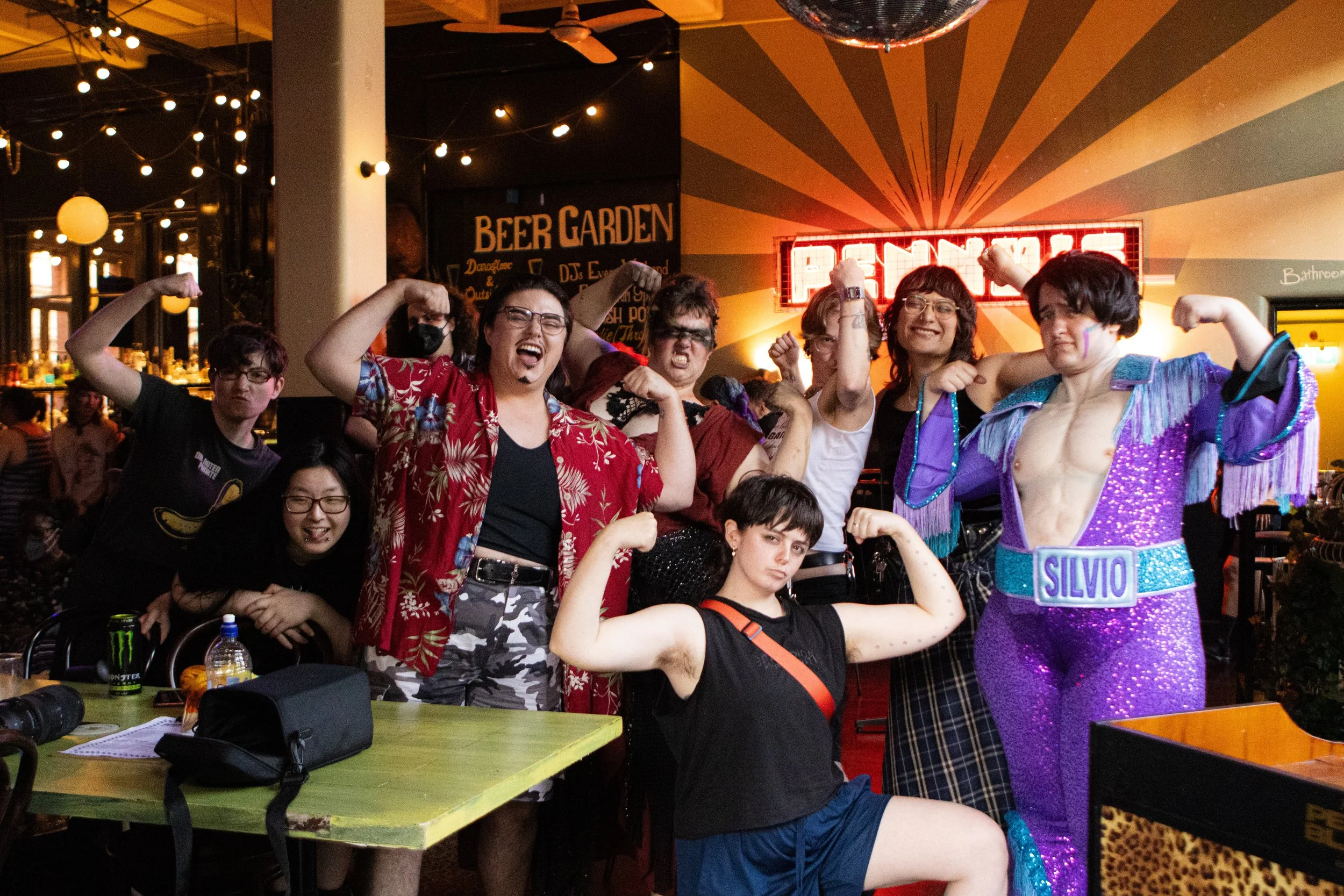 A group of people posing with flexed arms in a lively indoor space, with one person dressed in a sparkly purple costume with a 'Silvio' name tag, and others exhibiting playful expressions and gestures.