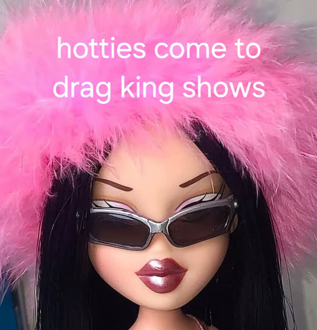 A doll with long black hair wearing large pink fuzzy hat, black sunglasses, and shiny lipstick. Pink text overlay reads 'hotties come to drag king shows'.