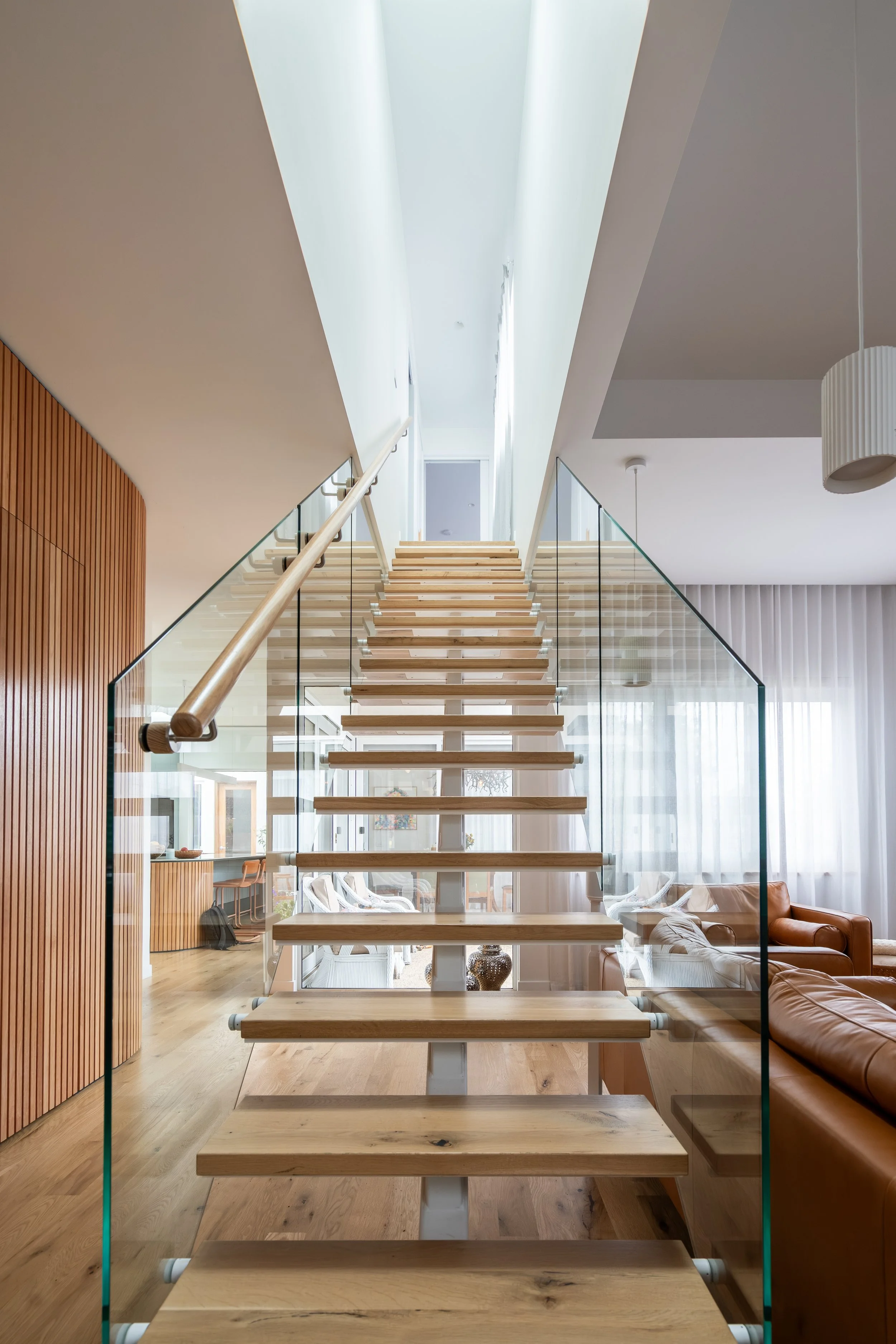 Interior view of a modern home featuring a floating wooden staircase with glass railings, leading to a second floor, in a well-lit living room with hardwood flooring and contemporary furniture.