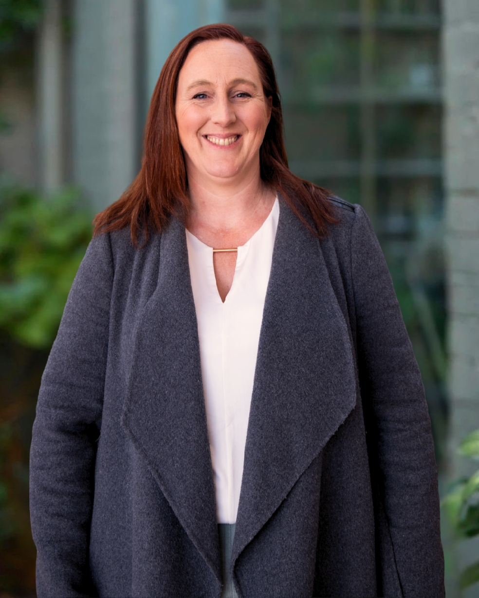 Anita Sweeney, Building and Interior Designer, Building Evolution. A smiling woman with shoulder-length red hair, wearing a dark gray coat over a white blouse, standing outdoors with a blurred background of greenery and structures.