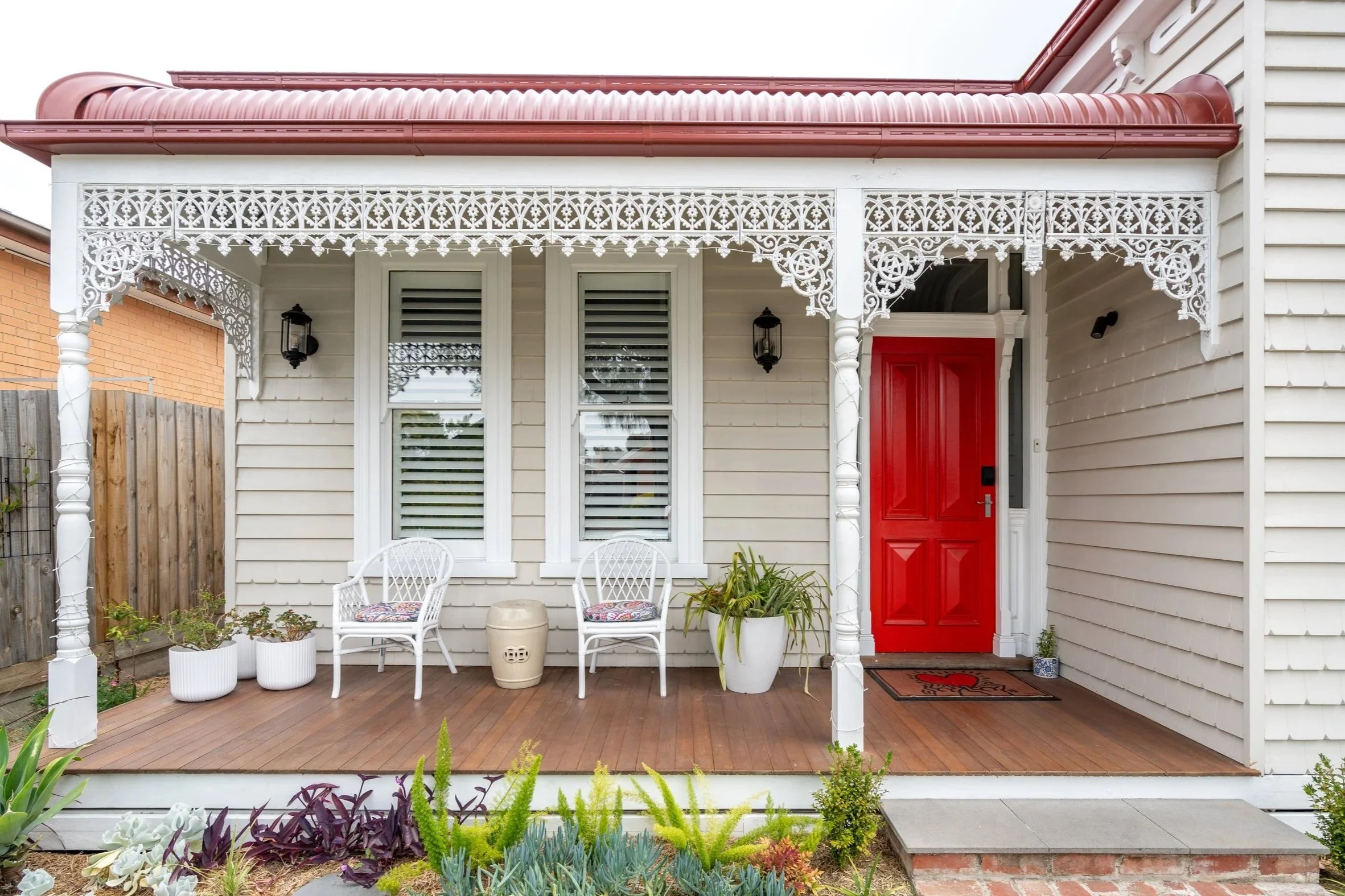 Front porch of a house with a red door, white trim, and decorative white lattice. Two white chairs with patterned cushions, potted plants, and a welcome mat are on the wooden porch. A small set of steps leads to a concrete and brick pathway.