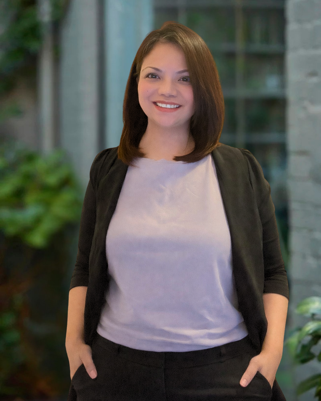 Lily Magpantay, Operations Coordinator, Building Evolution. Woman with brown hair smiling, wearing a light-colored top and a black jacket, standing outdoors in front of a building with large windows and plants.