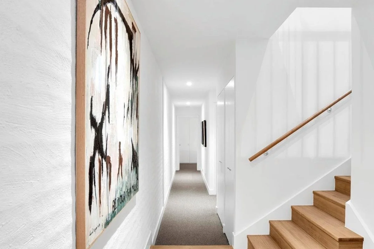 A clean, white hallway with framed artwork on the walls, carpeted floor, and wooden stairs with a handrail leading upward on the right side by Building Evolution.