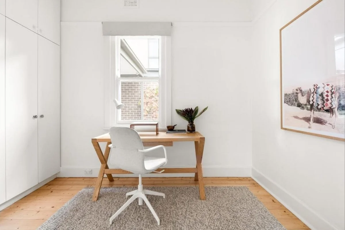 A minimalist home office with a wooden desk, white office chair, and a window with brick building view by Building Evolution.