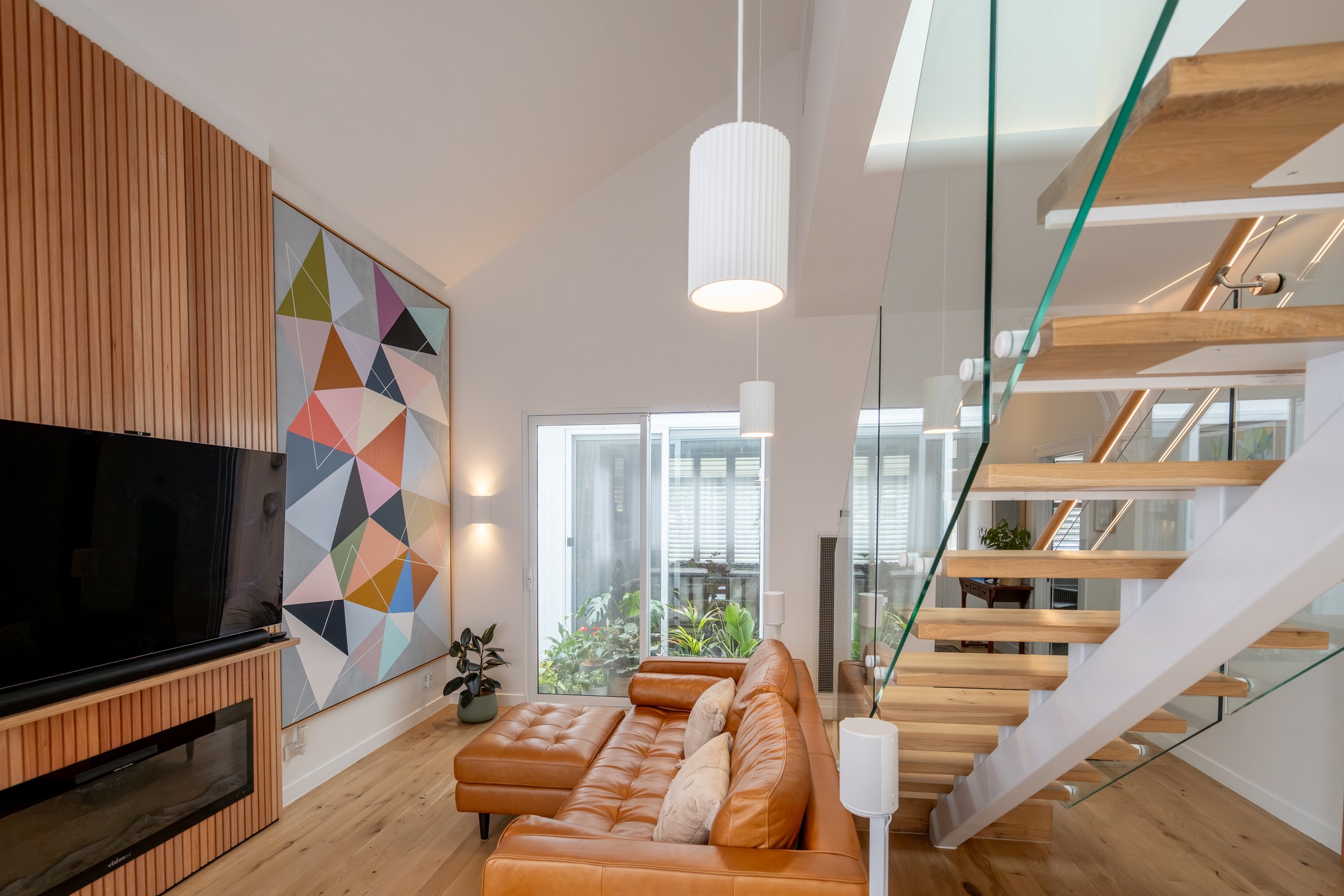 Living room with leather sectional sofa, colorful geometric wall art, wooden staircase with glass railing, and large window with plants outside.