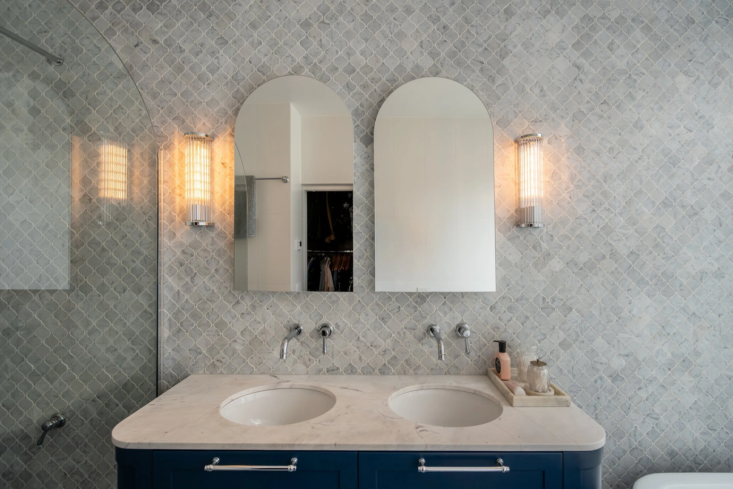 A bathroom vanity with a marble countertop, two undermount sinks, and a blue cabinet. Two mirrors are mounted above the sinks, flanked by wall sconces. A tray with toiletries is on the right side of the countertop.