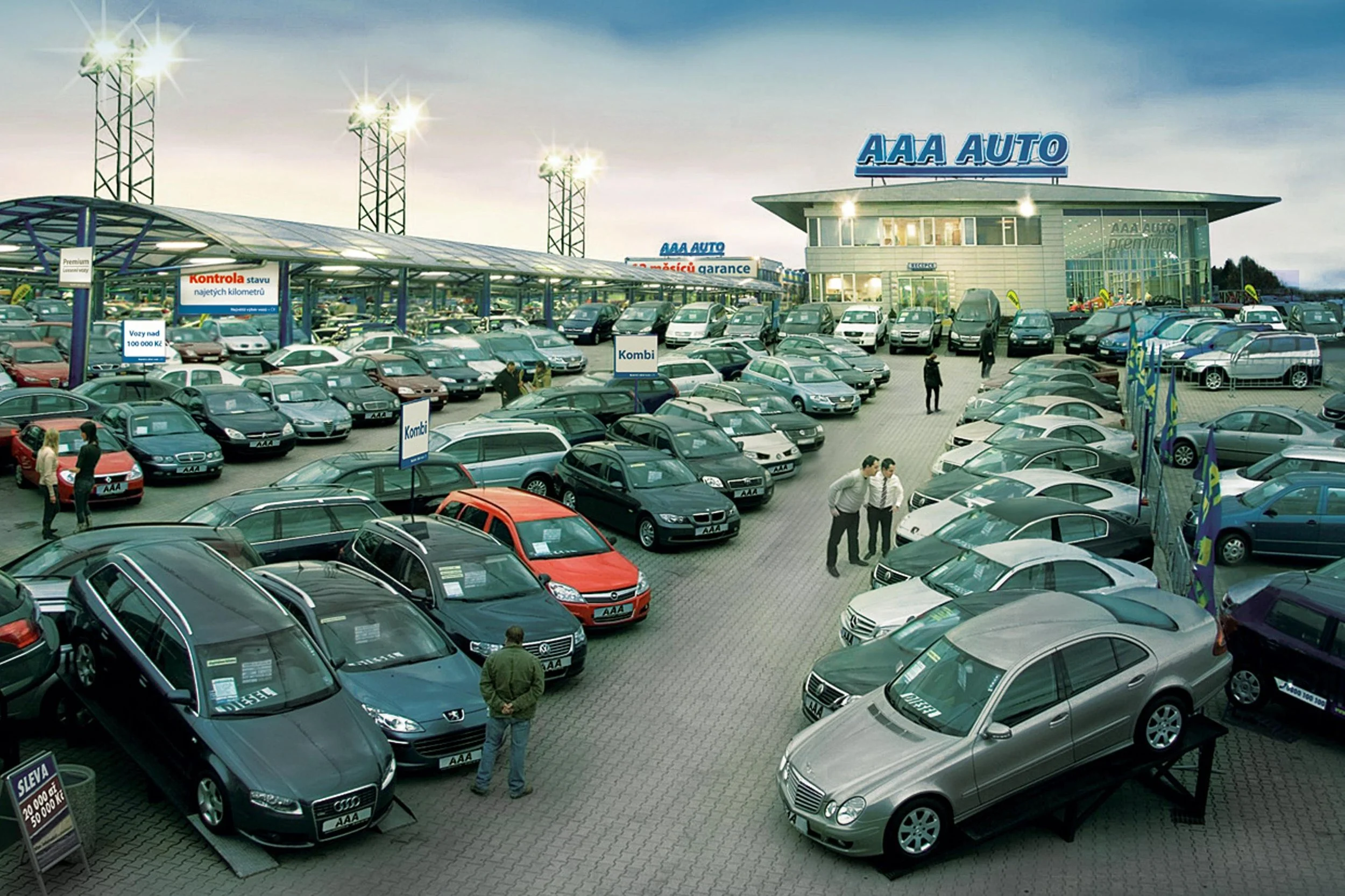 Large car dealership lot filled with various new and used cars, with a building labeled 'AAA AUTO' in the background and people walking around inspecting vehicles.