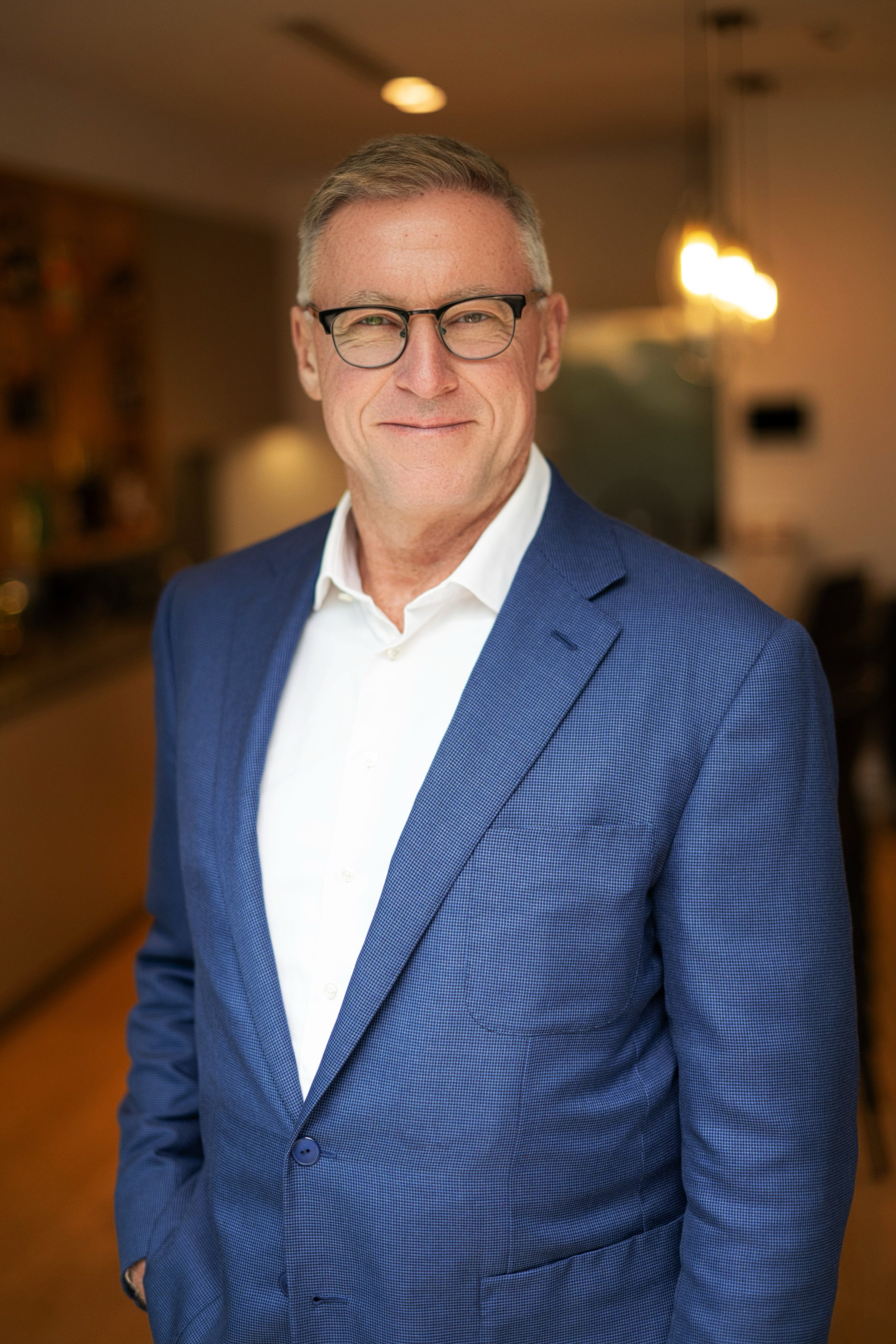 A man with gray hair, glasses, wearing a white shirt and a blue suit, smiling at the camera indoors.