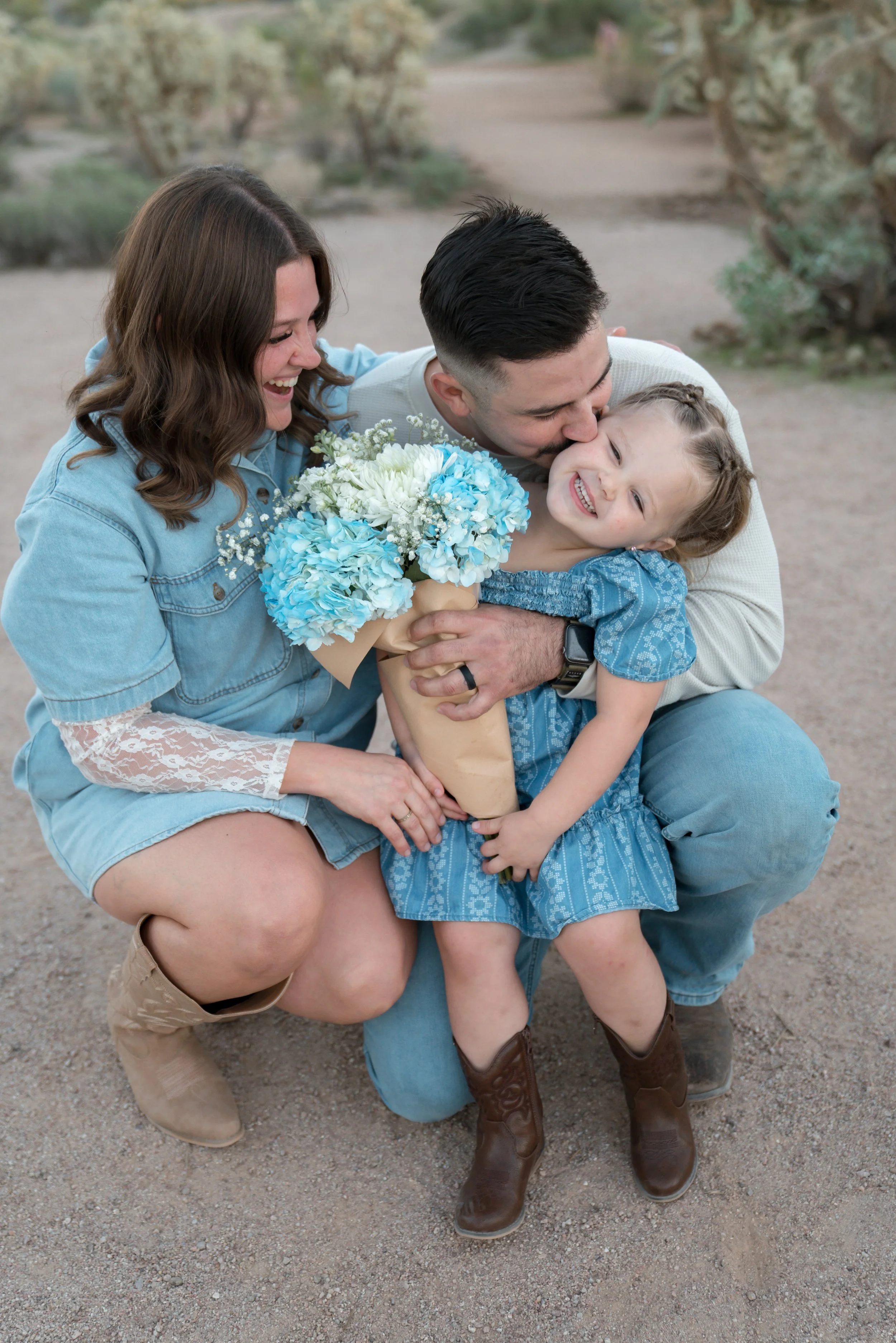 A happy family of three, a mother, father, and young daughter, sharing a joyful moment outdoors on a dirt path with desert plants in the background. The mother and father are embracing their smiling daughter, who is holding a bouquet of blue and white flowers and wearing a blue dress and brown cowboy boots.