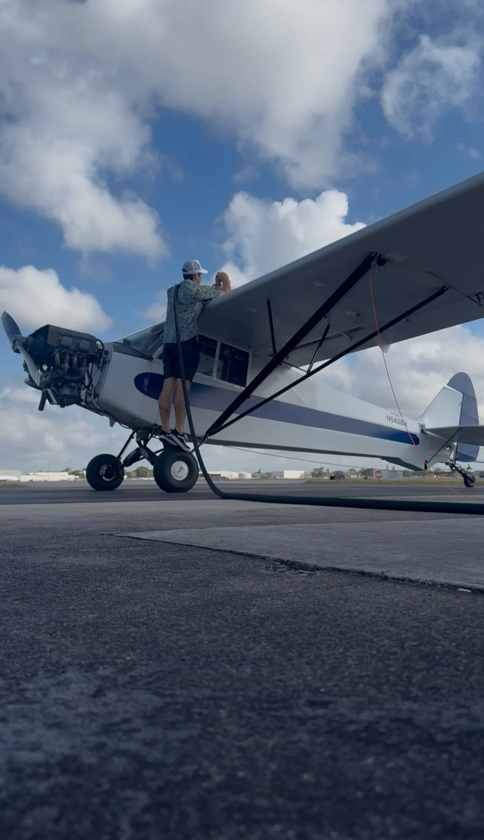 A man filling a small propeller airplane with fuel on the tarmac, under a partly cloudy sky.