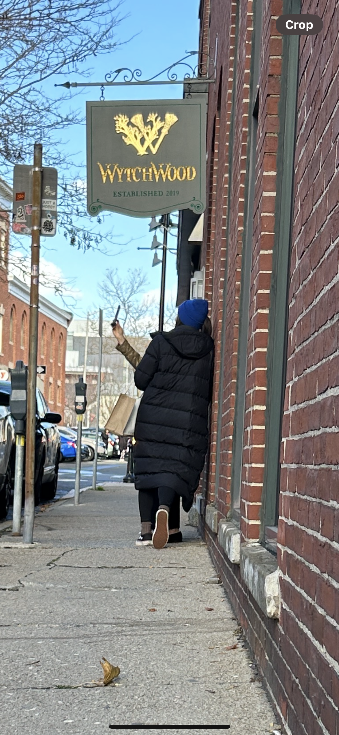 Person in a black winter coat and blue hat walking past a brick building with a sign for Witchwood established in 2019, while taking a photo or video with a smartphone.