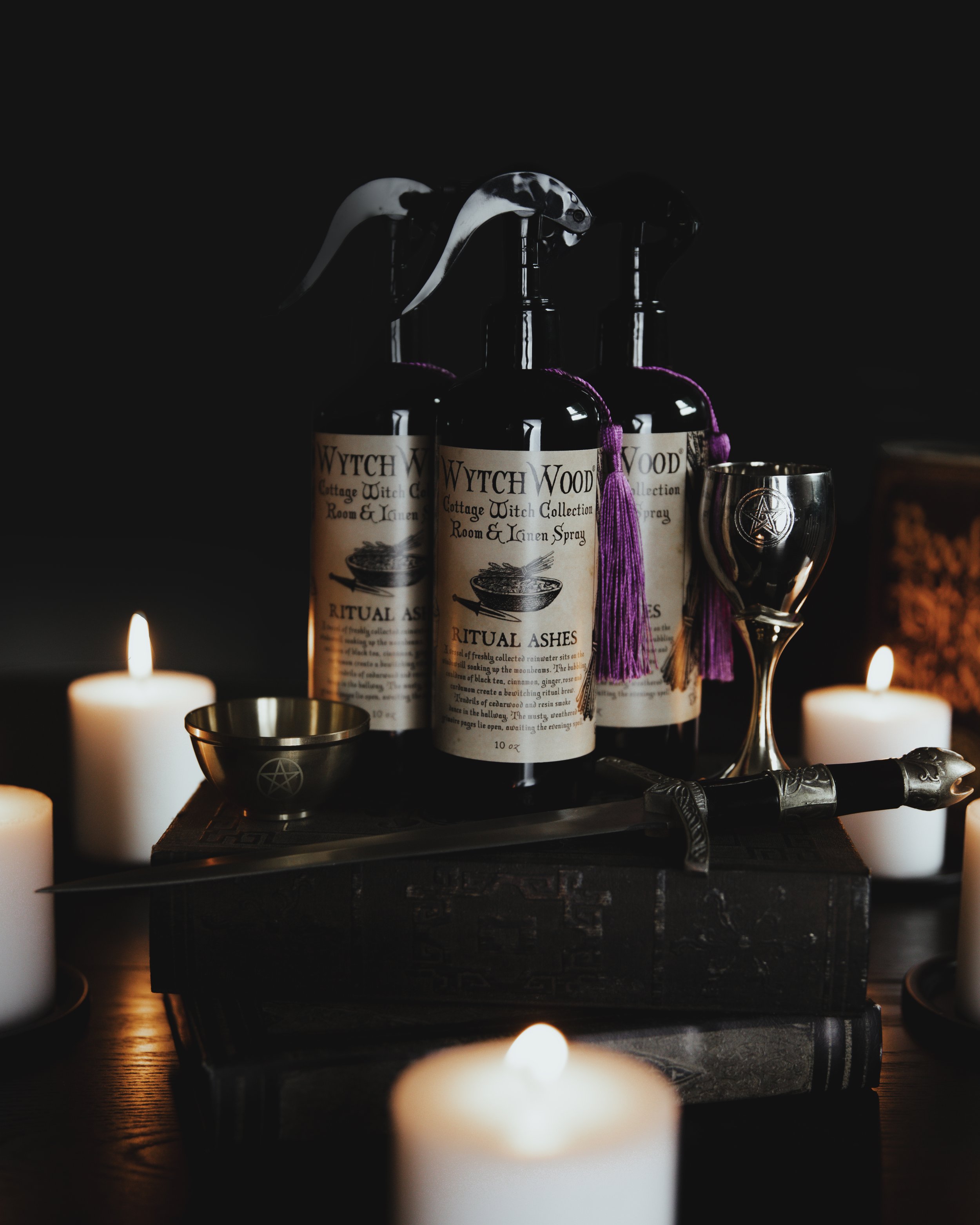Dark-lit scene with candles, bottles labeled 'Witch Wood' Ritual Ashes spray, a chalice, a small bowl with a pentagram, a ritual dagger, and books.