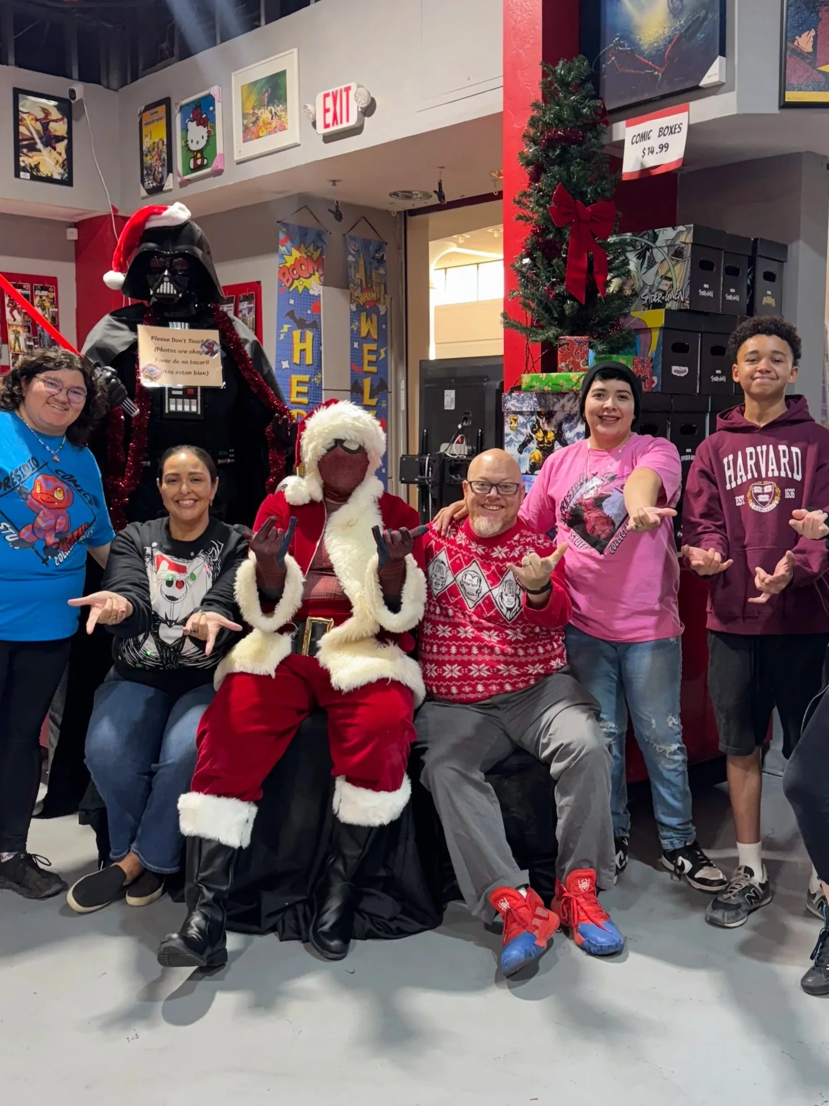 People posing with Santa, Santa's helper, and a Darth Vader figure wearing a Santa hat in a holiday-themed setting at a comic or game store.