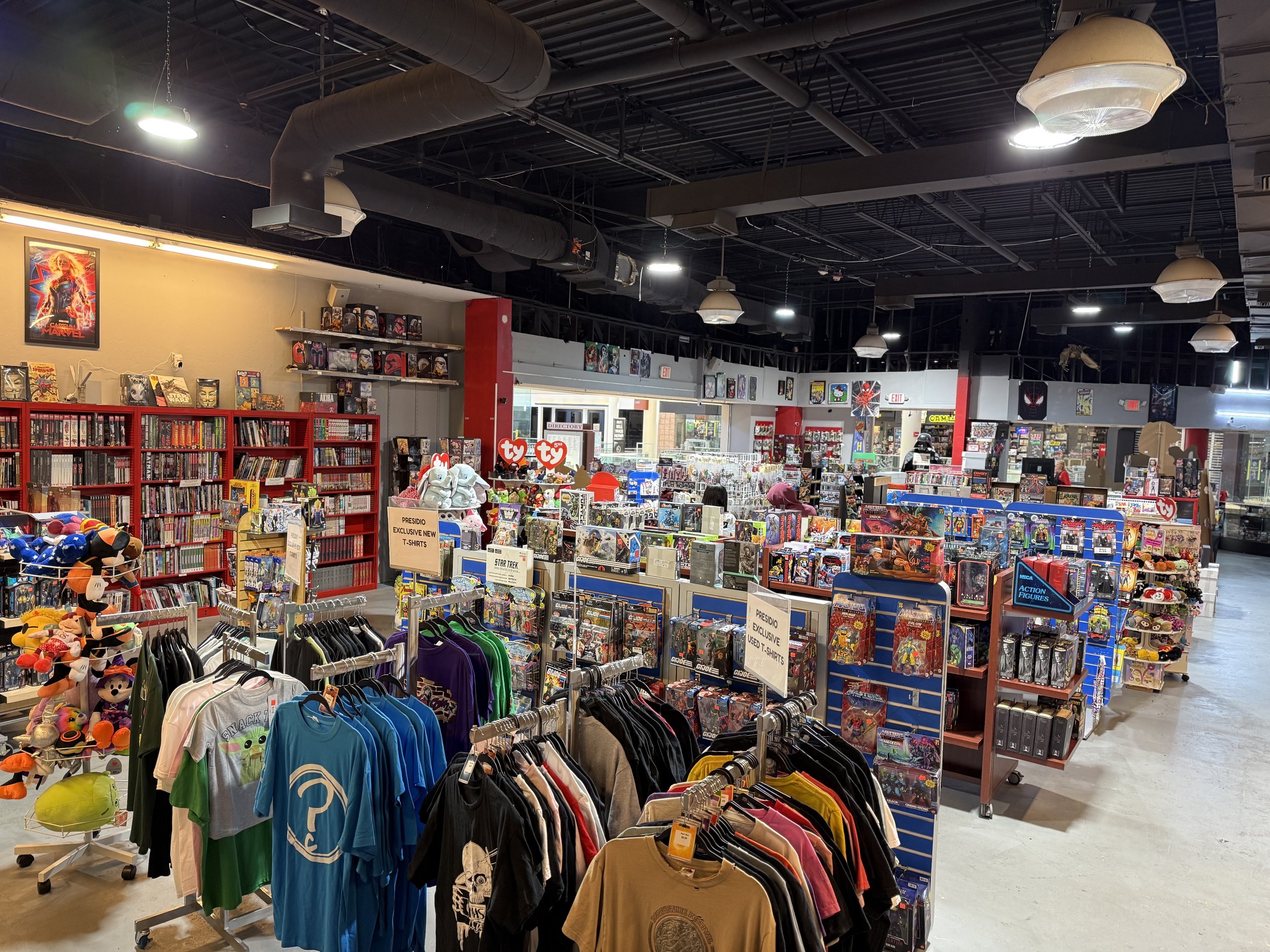 Interior of a retail store selling comic books, toys, clothing, and collectibles with shelves filled with merchandise, posters, and signage.