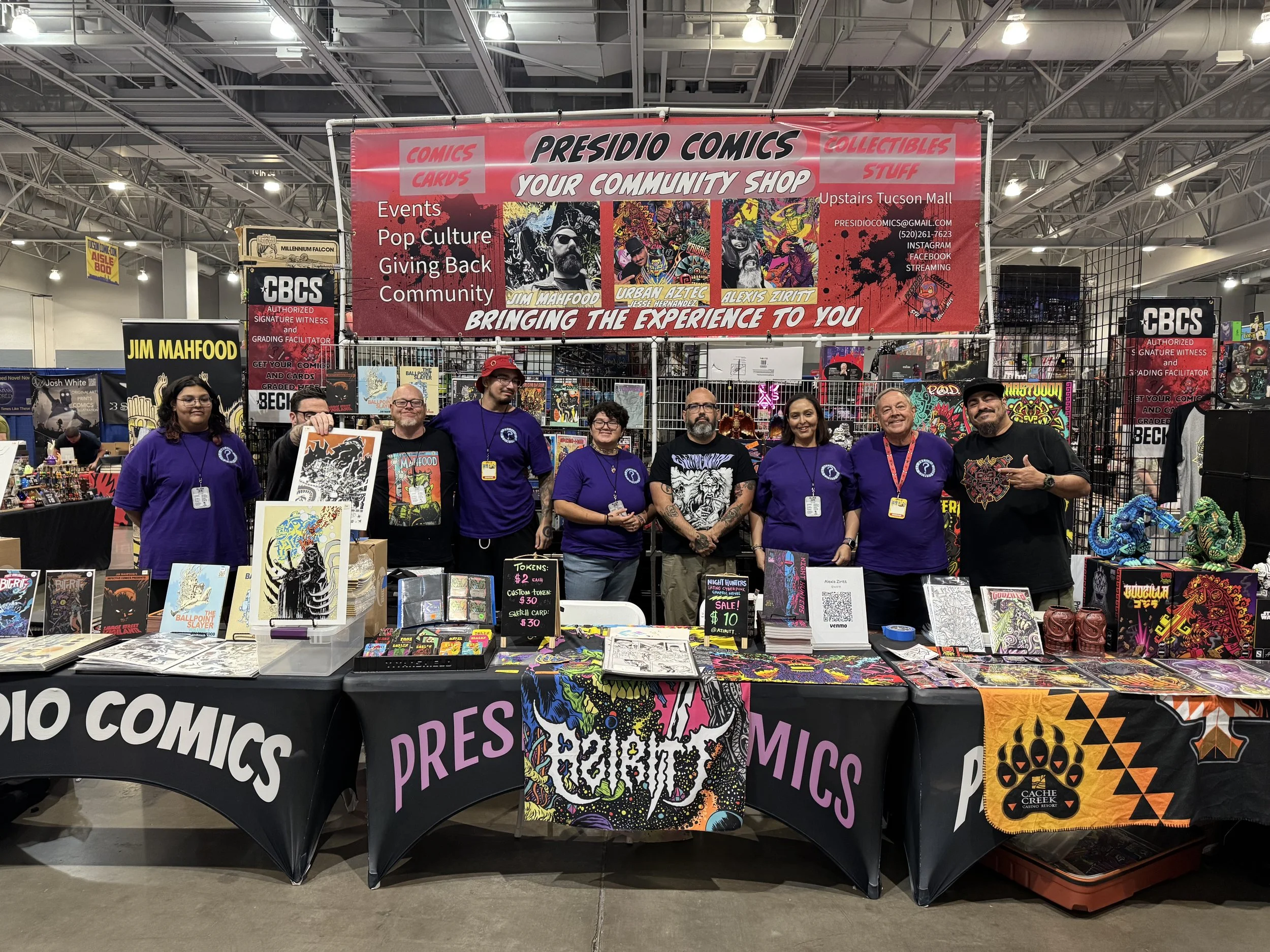Group of people standing behind a booth at a comic convention, displaying comics, art prints, and merchandise, with banners and signs for Presidio Comics and other vendors.
