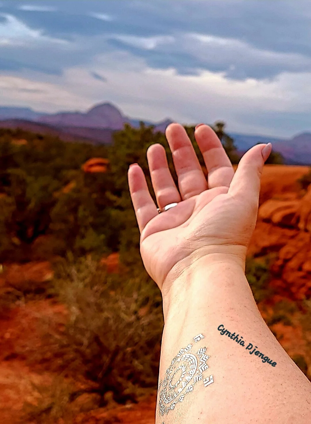 Cynthia's hand reaching out, raised against a backdrop of red rock formations in Sedona, AZ and a cloudy sky.