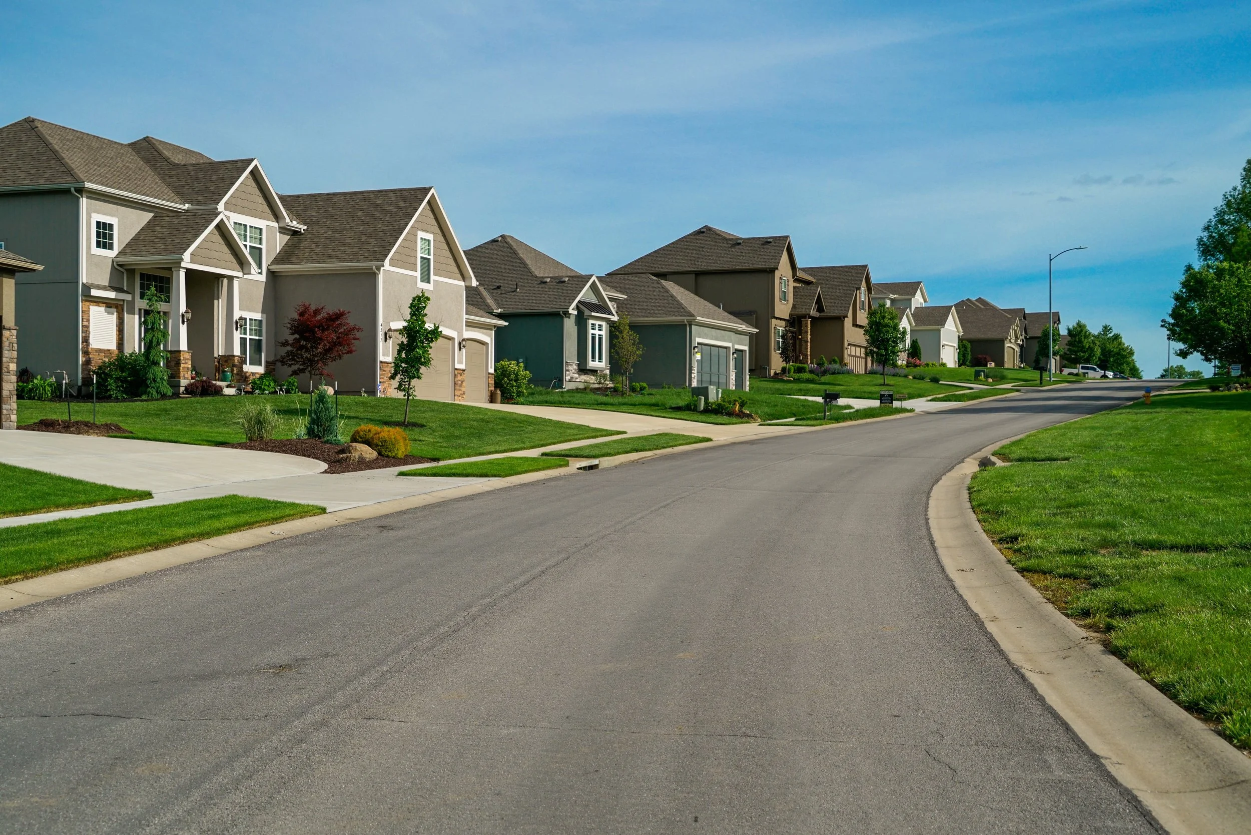 A suburban neighborhood street with well-maintained houses, green lawns, and landscaped gardens, under a partly cloudy sky.