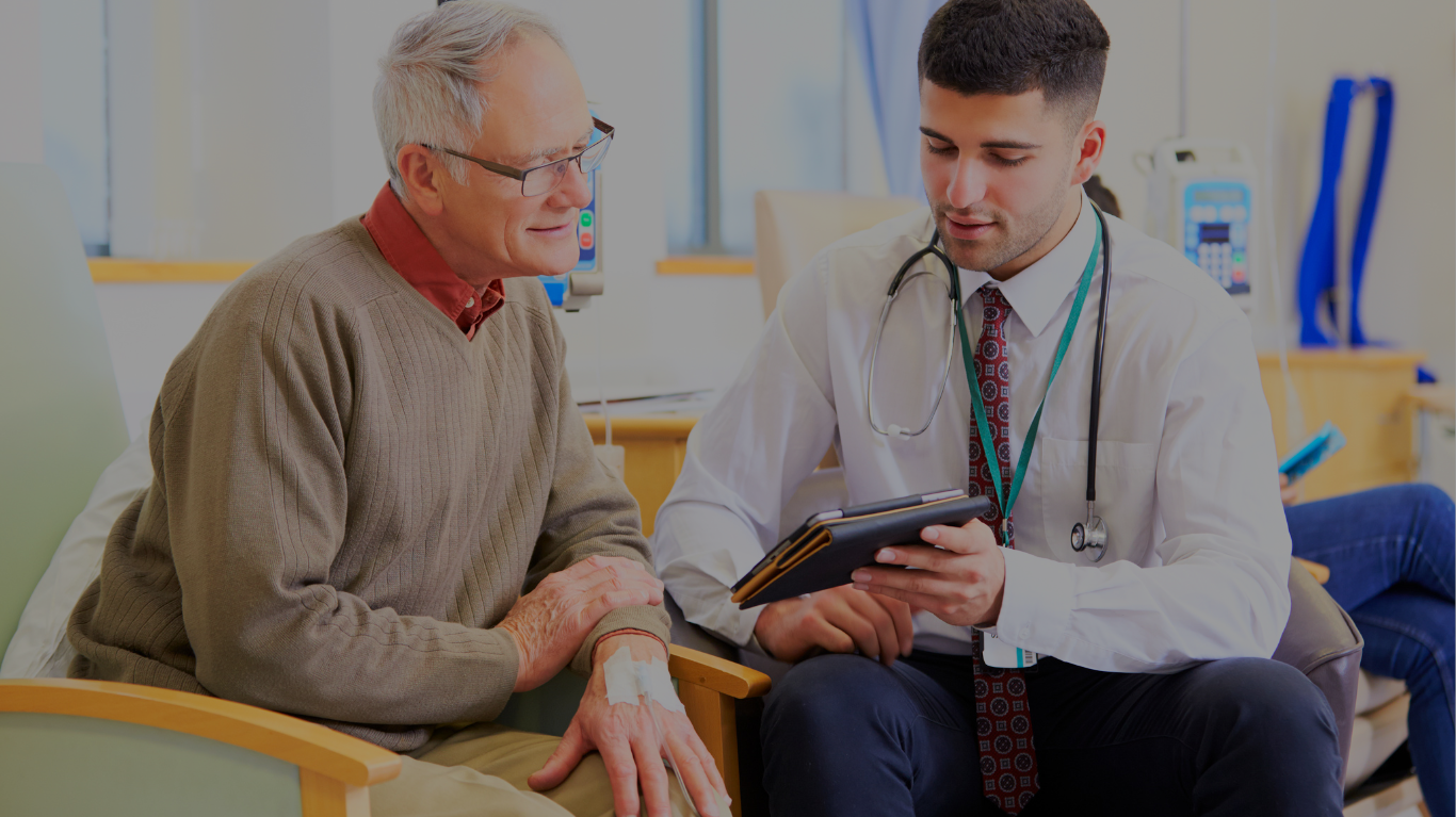 A doctor consulting with an elderly male patient in a hospital room. The patient is sitting on a bed with an IV in his arm, while the doctor is holding a tablet and discussing something with him.