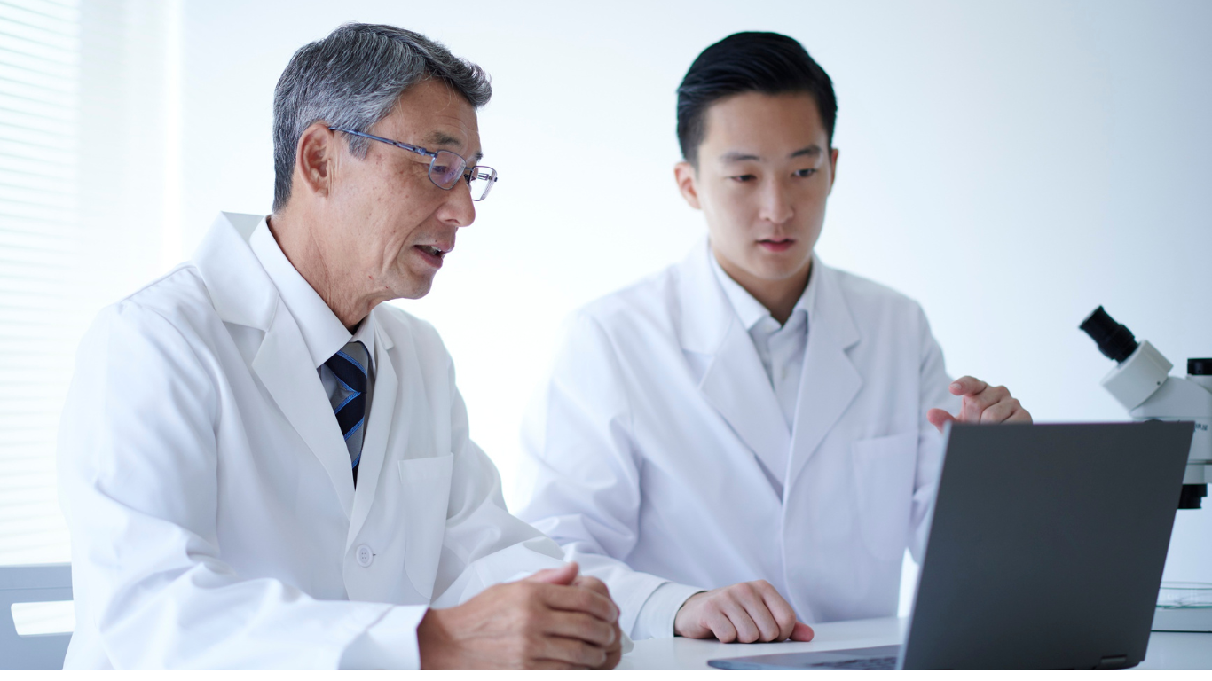 Two scientists in lab coats working together with a laptop, one is older with glasses, the other younger, in a laboratory setting.