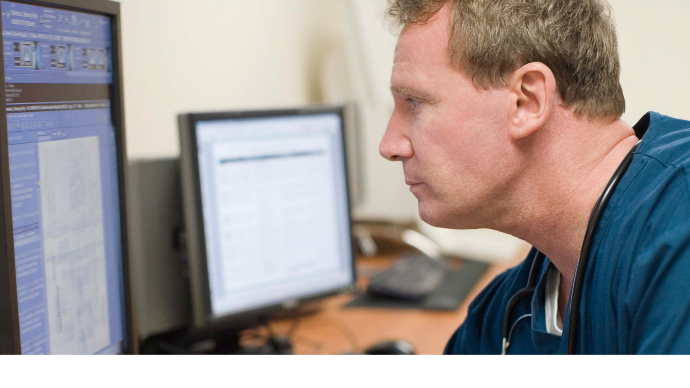 A male healthcare professional with a stethoscope around his neck working on multiple computer monitors in an office or medical setting.