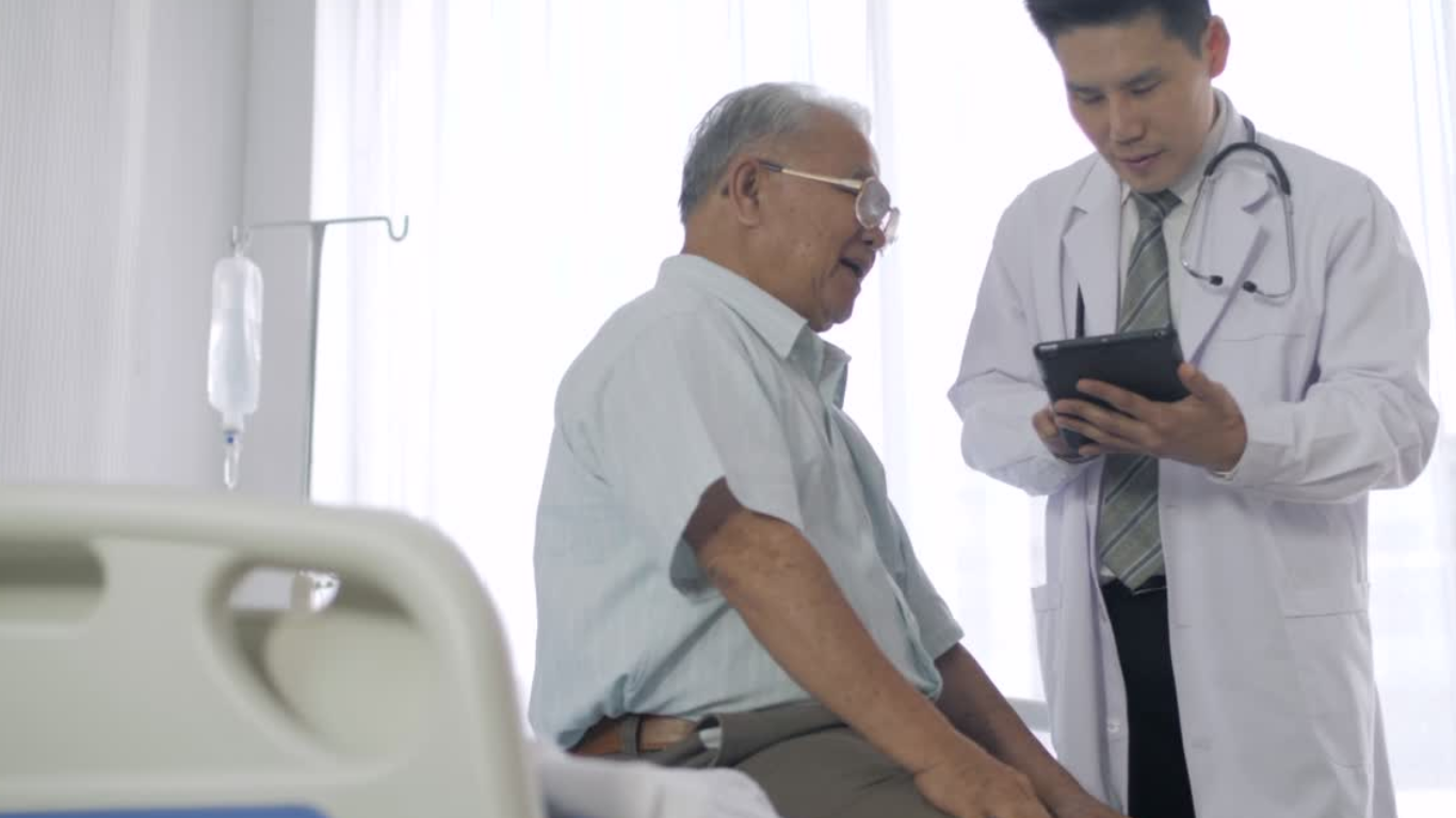 A doctor showing a tablet to an elderly patient in a hospital room with an IV drip in the background.