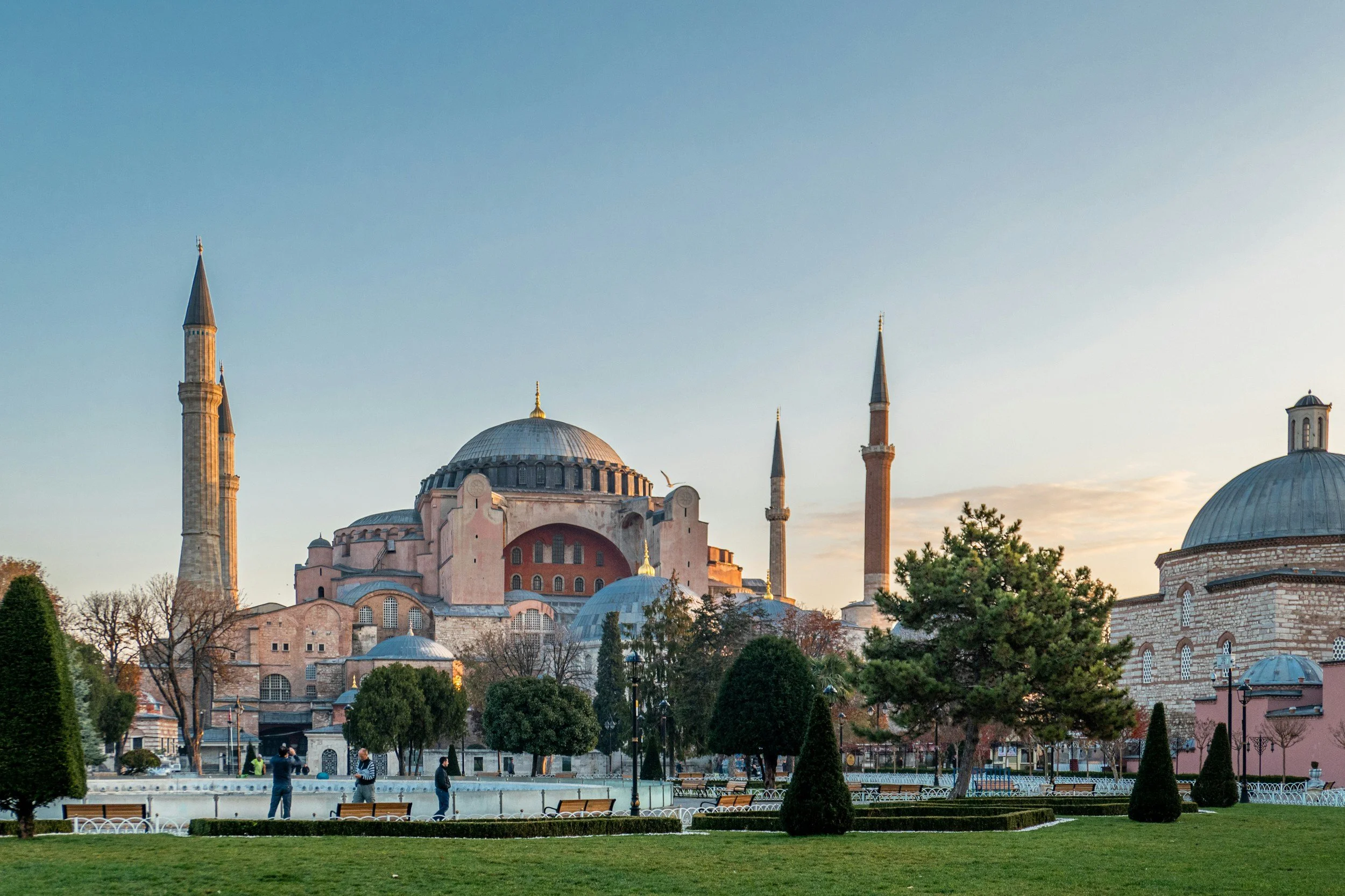 View of Hagia Sophia with domes and minarets, green park with trees and benches in the foreground at sunset.